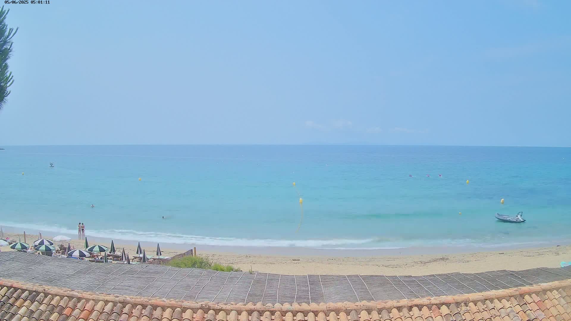A sandy beach with several umbrellas and a few people is visible, overlooking a calm, light-blue ocean under a clear sky.