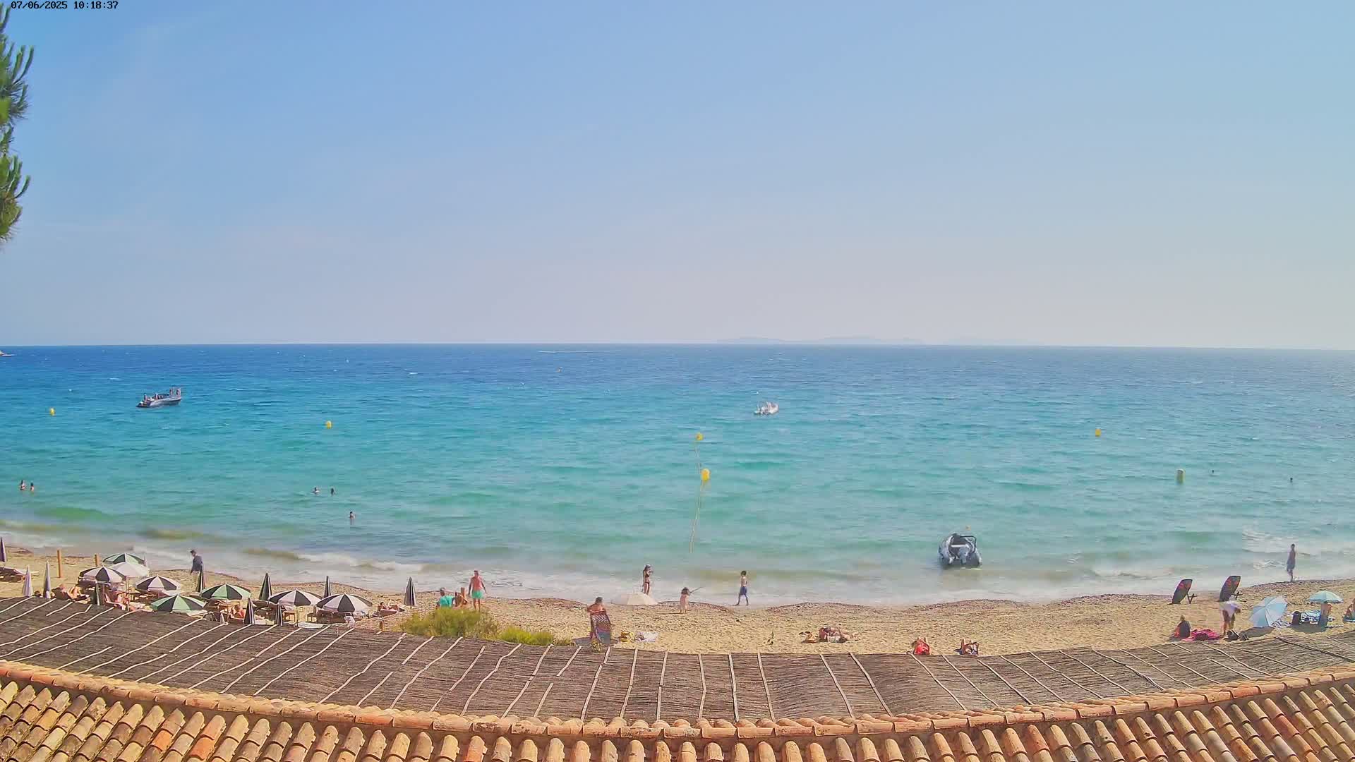 A sandy beach with scattered people and umbrellas is visible below a thatched roofline, overlooking a calm, turquoise sea on a sunny day.