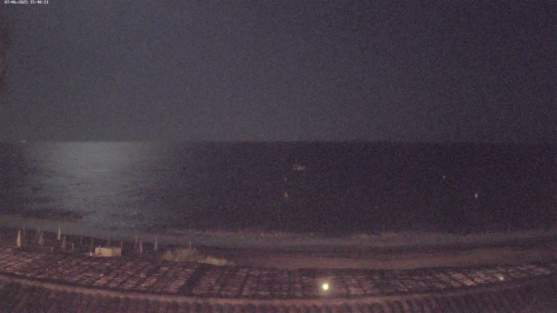 A dark, nighttime view shows a calm, dark ocean under a cloudy sky, with a shoreline featuring a flat, dark expanse that appears to have some sort of stacked objects.