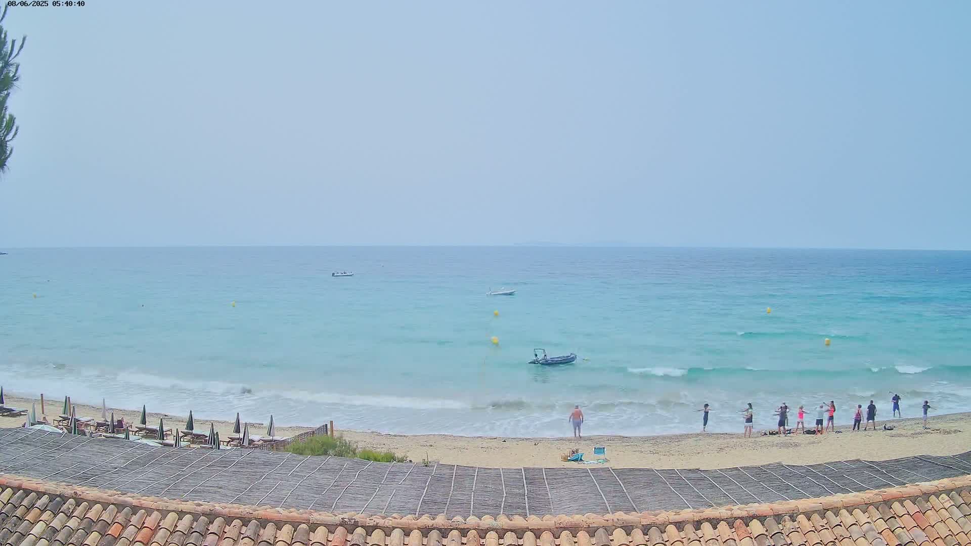 A sandy beach with calm, light-blue water and several people standing on the shore under a hazy sky.