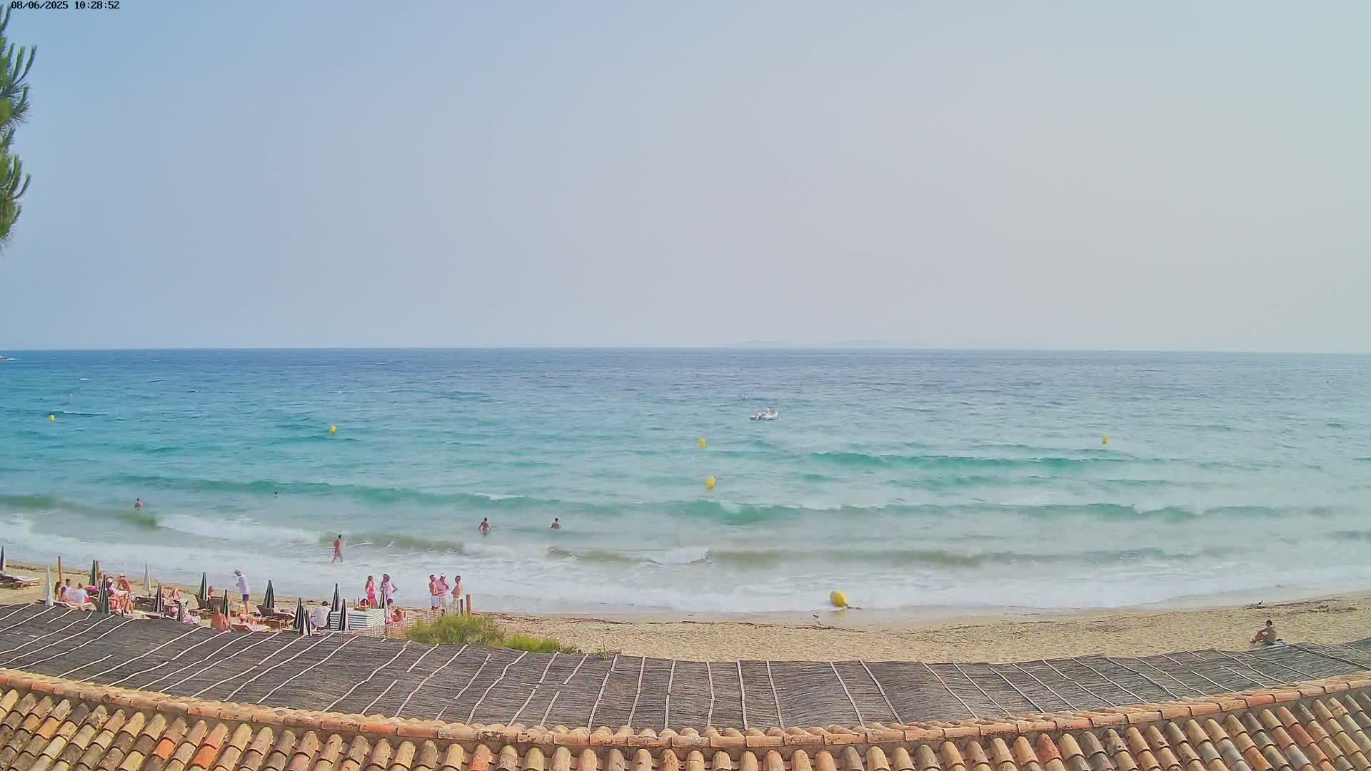 A sandy beach with people swimming and sunbathing, viewed from a rooftop with a tiled roof, under a mostly clear sky and gentle waves on a turquoise sea.