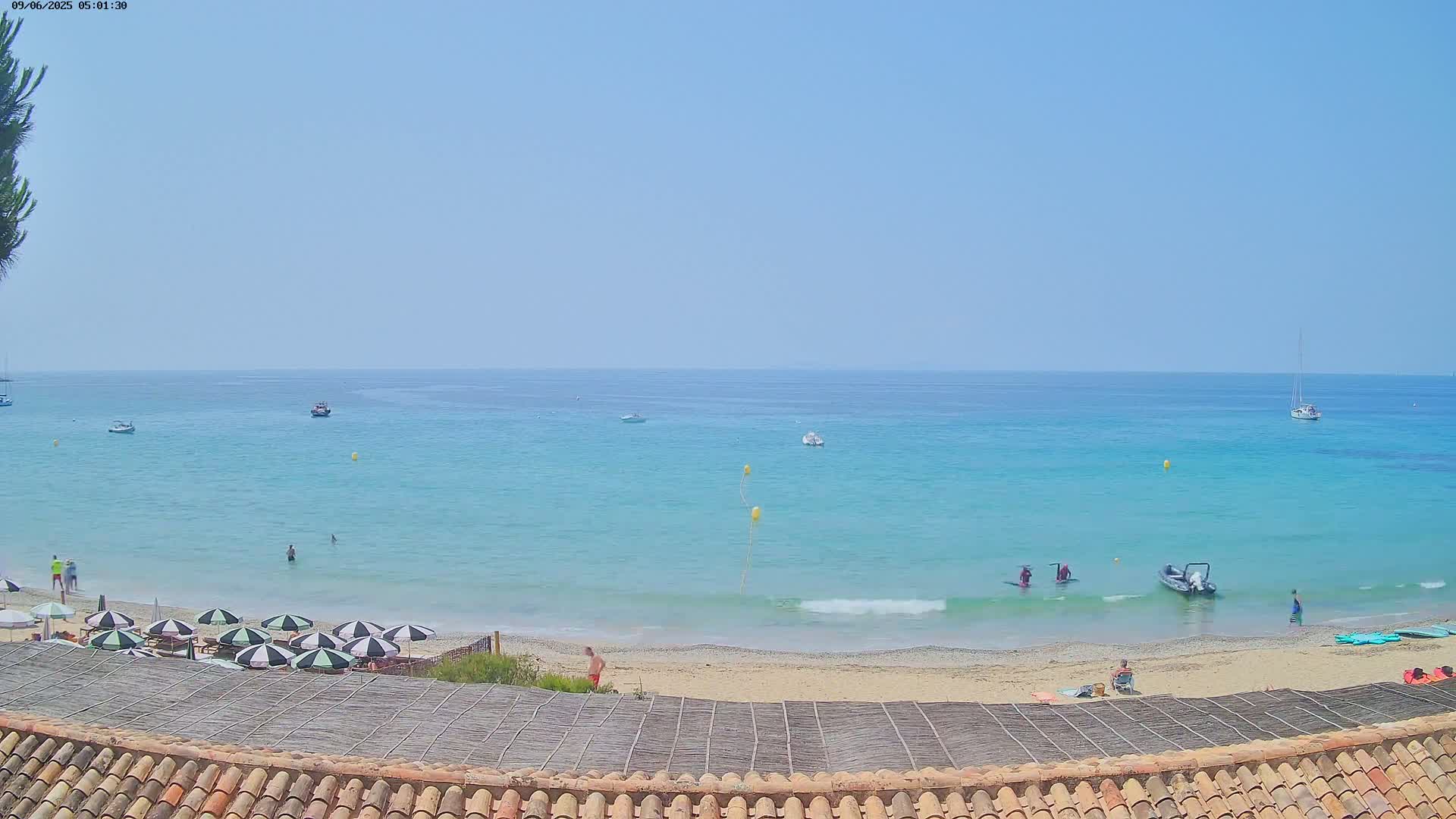 A sandy beach with turquoise water, several boats, and a few people swimming and sunbathing under a clear sky.