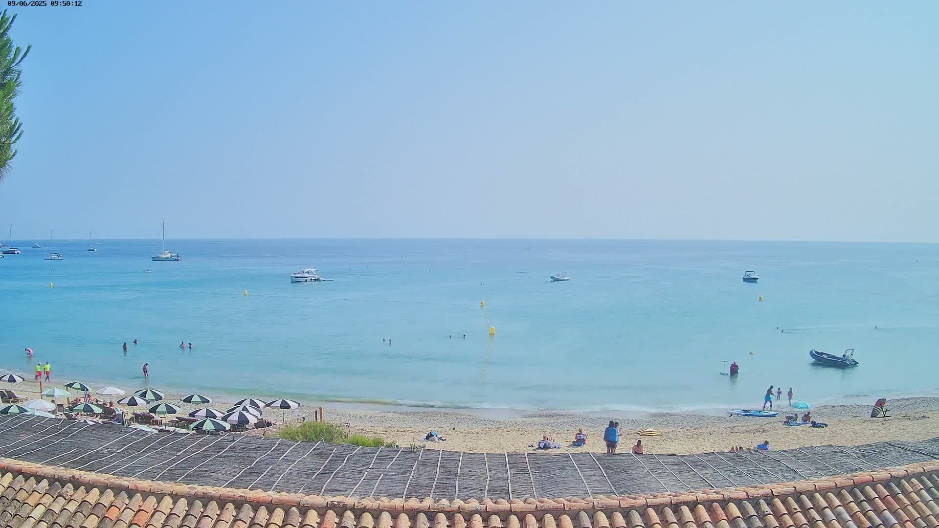 A beach scene on a sunny day shows a calm, light-blue sea with several boats and people swimming and sunbathing on the sandy shore.