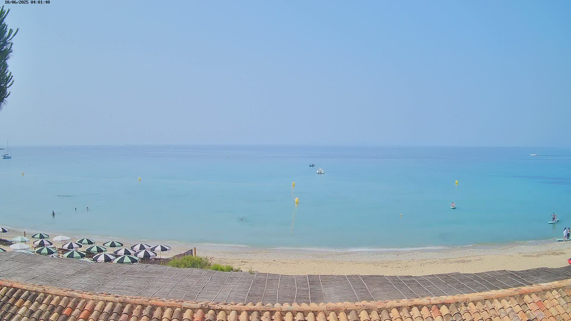 A sandy beach with teal water and several striped umbrellas is viewed from above on a clear, sunny day.