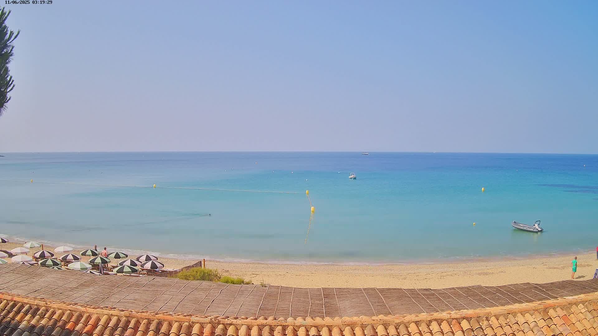 A sandy beach with striped umbrellas and a few people, overlooks a calm, light-blue sea on a clear day.