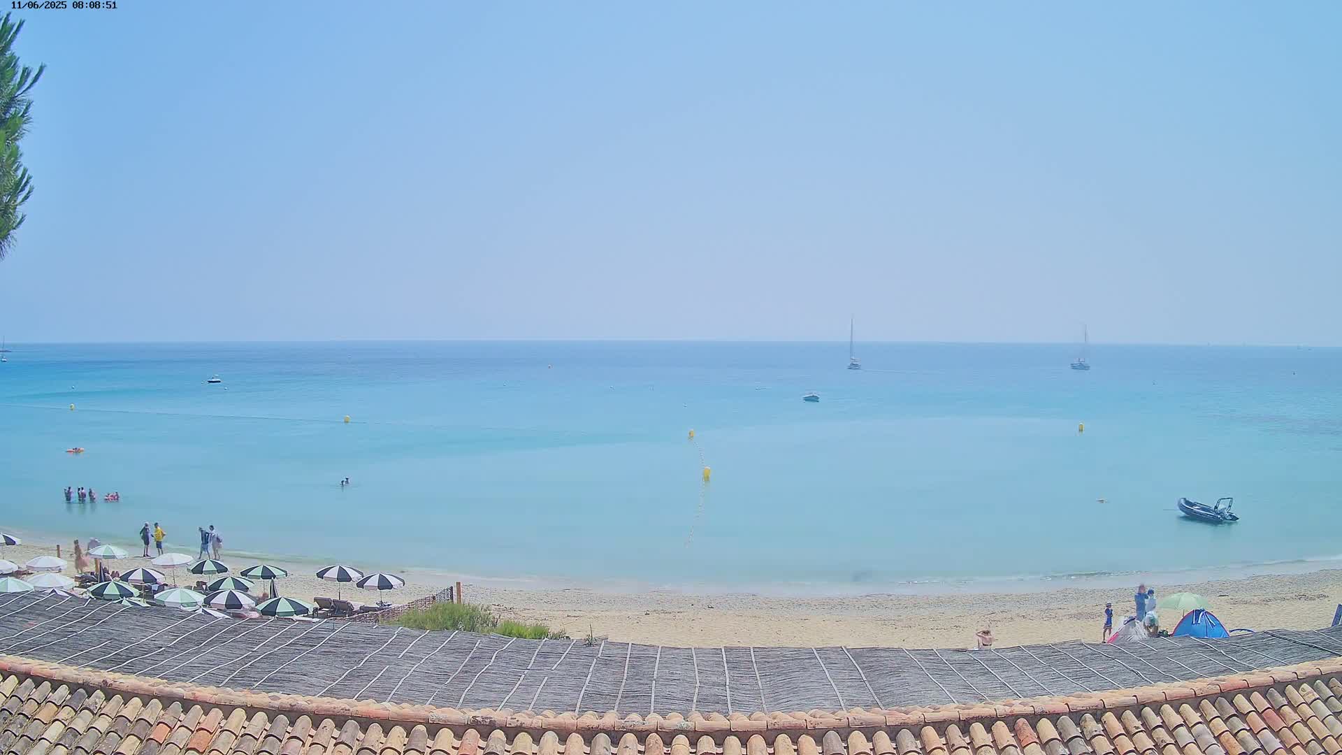 A sandy beach with several people and umbrellas is visible, overlooking a calm, light-blue ocean with a few boats under a clear sky.