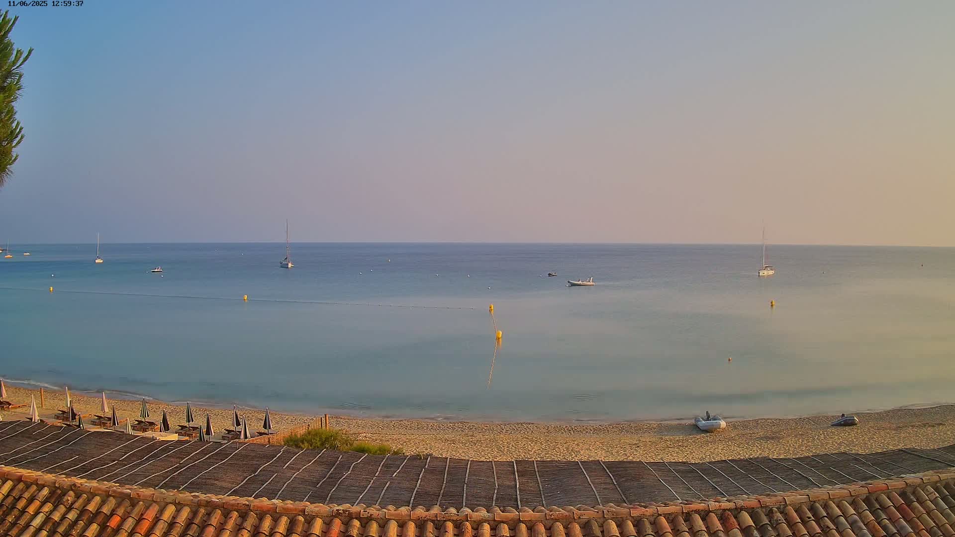 A sandy beach with several lounge chairs and umbrellas overlooks a calm, light-blue sea with several sailboats and small motorboats under a clear, pale sky.