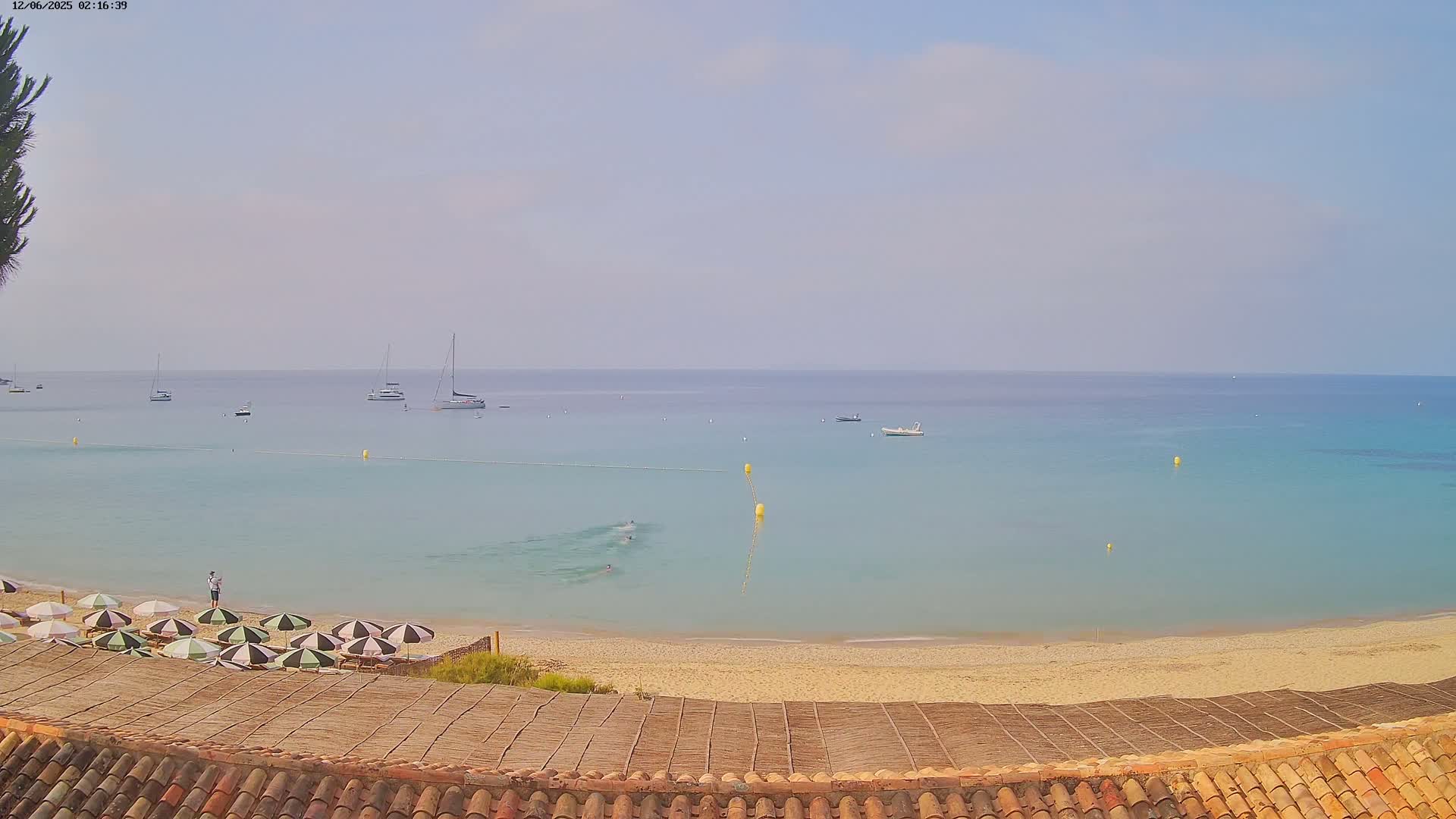 A sandy beach with several striped umbrellas and a calm, clear turquoise sea dotted with sailboats under a hazy sky.