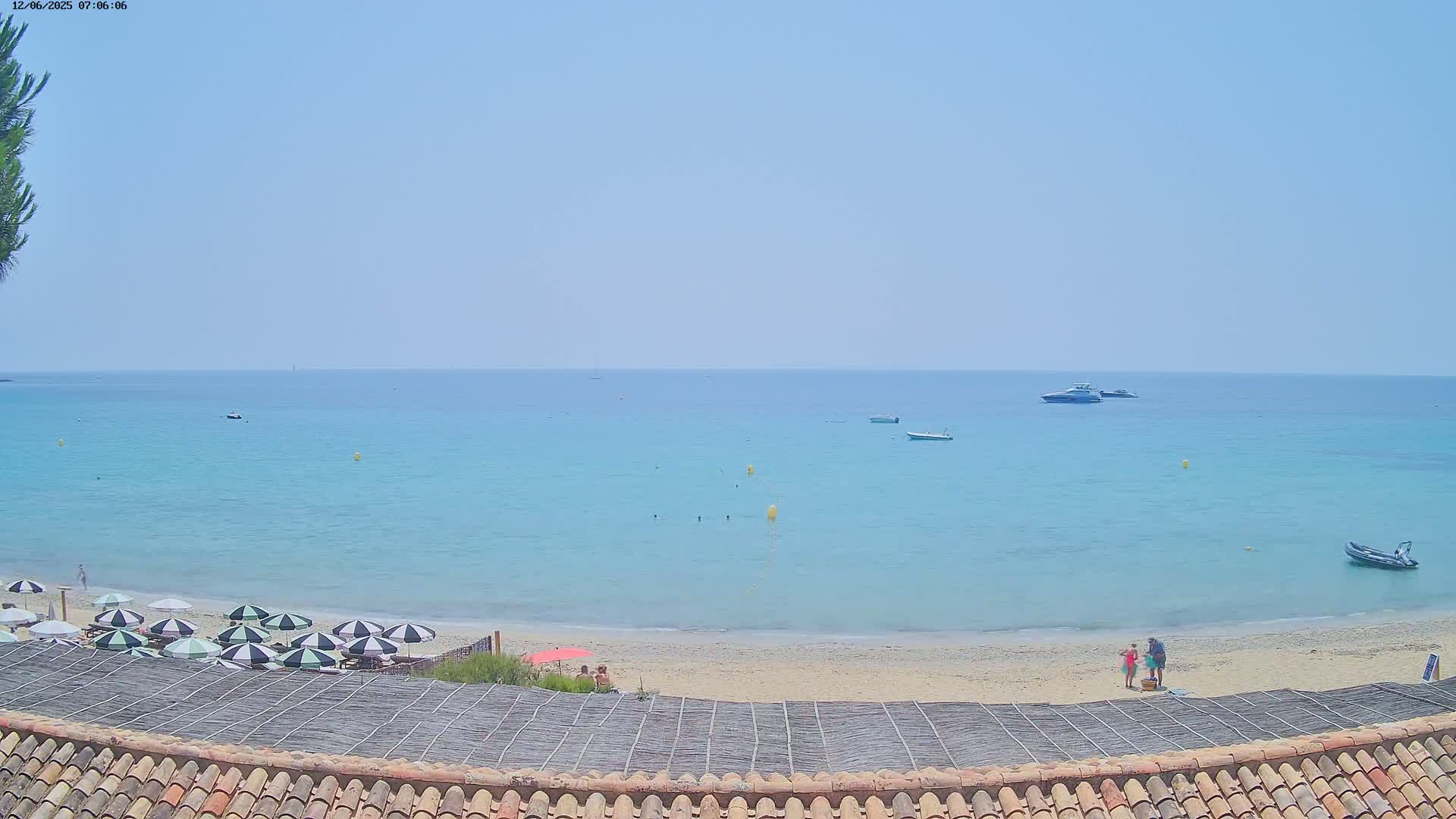A sandy beach with several striped umbrellas and a few people, overlooks a calm, light-blue ocean with several small boats on a clear, sunny day.