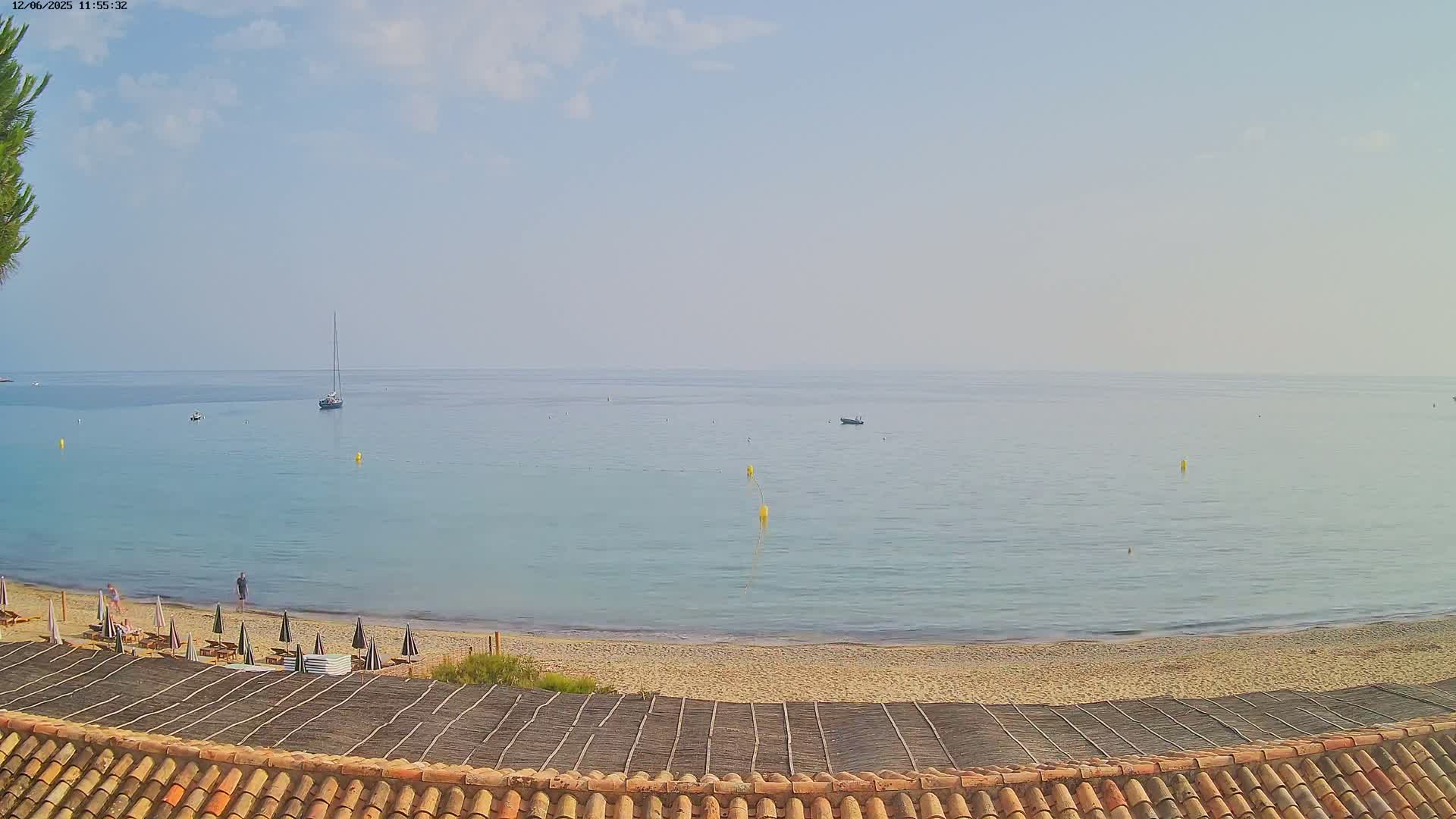 A sandy beach with a few people and umbrellas overlooks a calm, light-blue sea with a sailboat and small boats in the distance under a mostly clear sky.