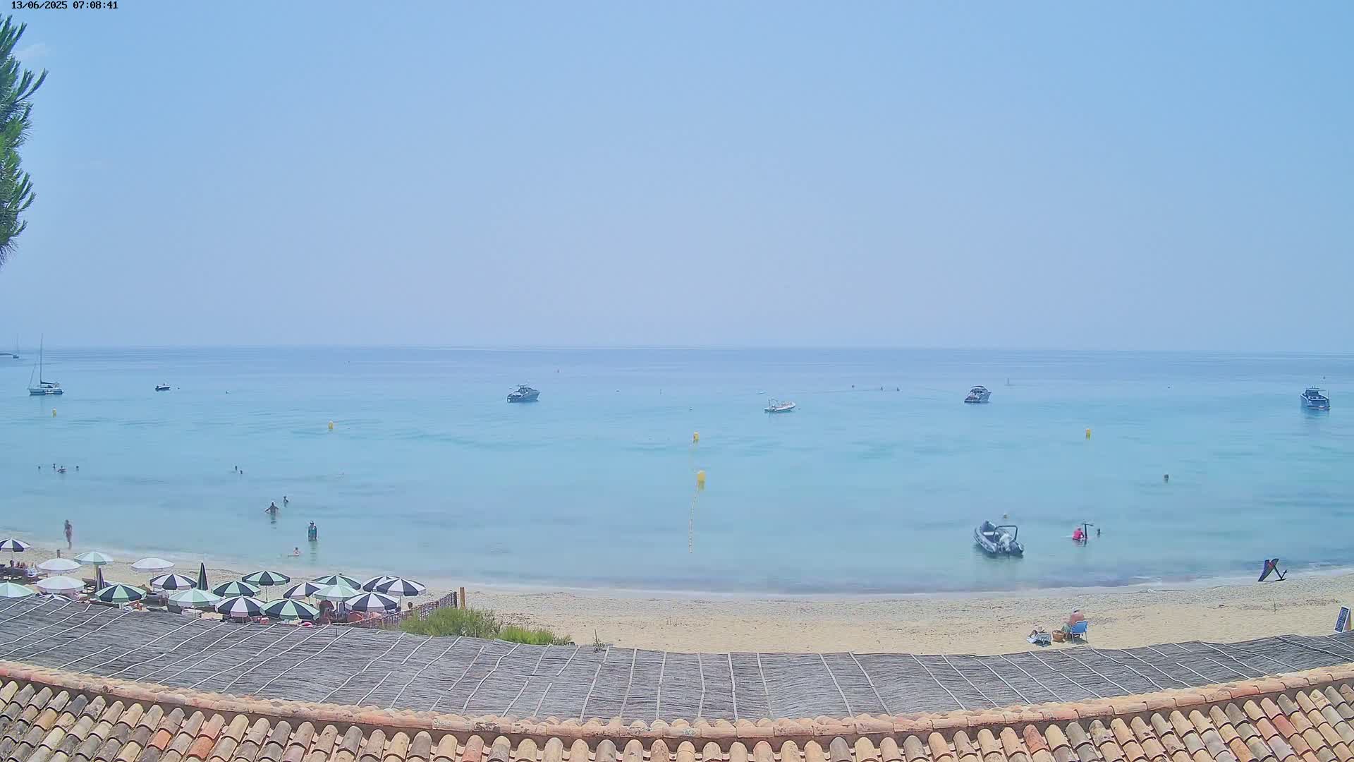 A sandy beach with several people and umbrellas is situated beside a calm, light-blue ocean with several boats under a clear, bright sky.