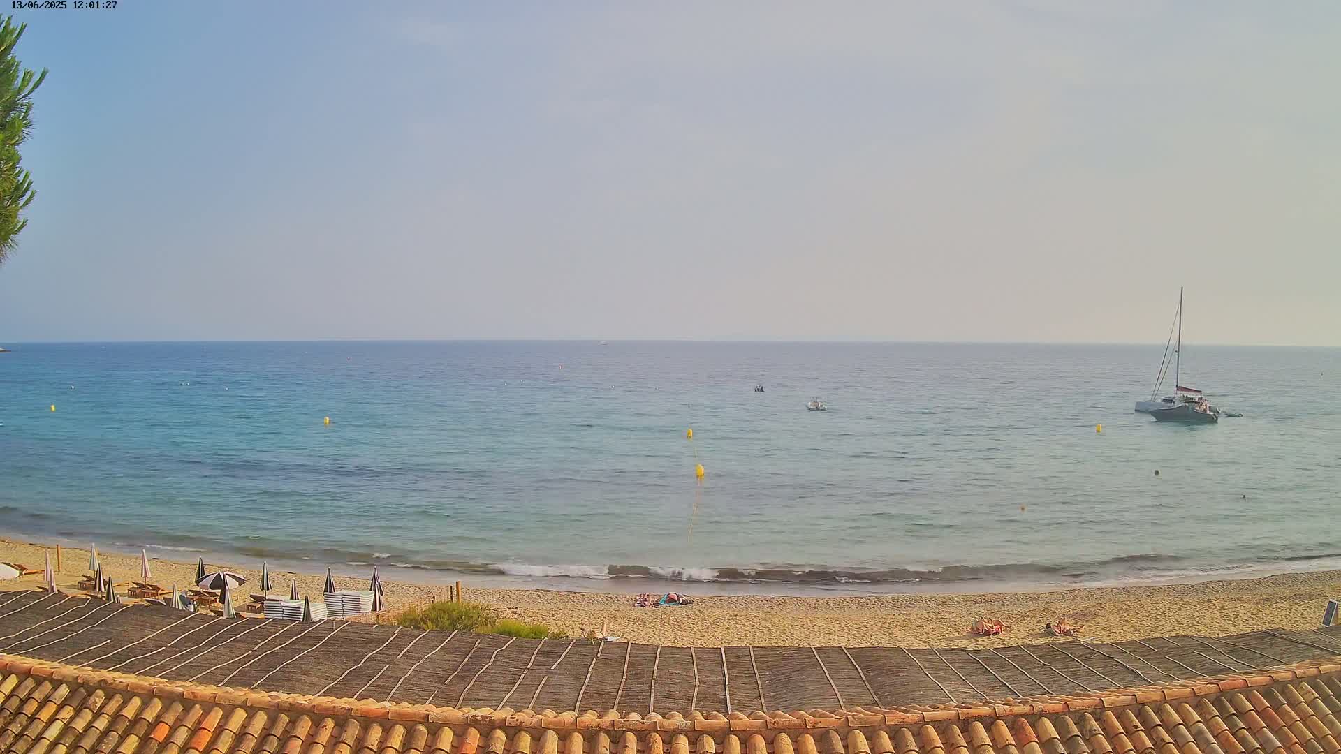 A sandy beach with a few sunbathers and beach umbrellas is visible from behind a brown thatched roof, overlooking a calm, clear ocean with several boats and yellow buoys.