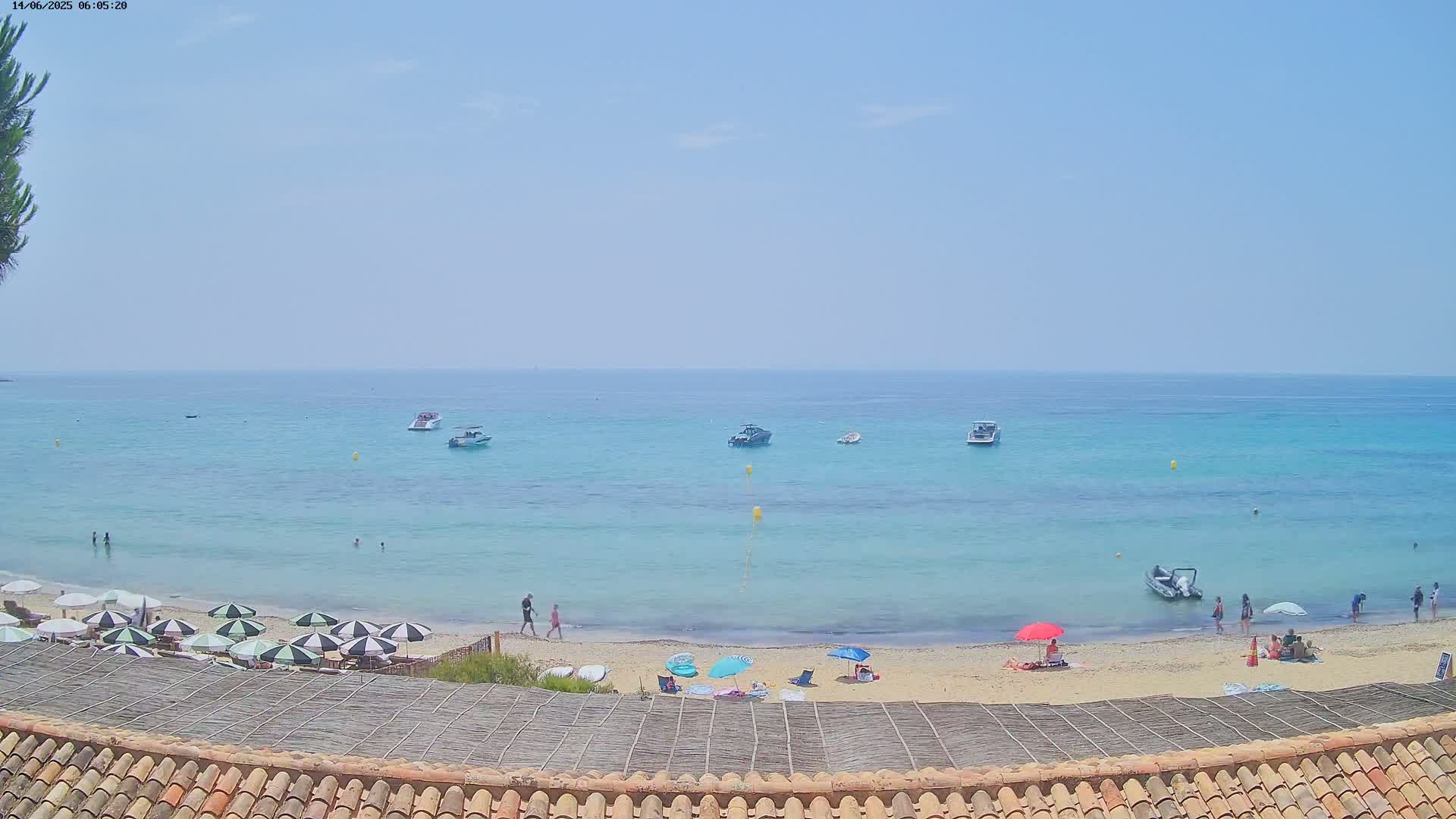 A sandy beach with several people and umbrellas is visible, overlooking a calm, light-blue sea with several small boats under a hazy, sunny sky.