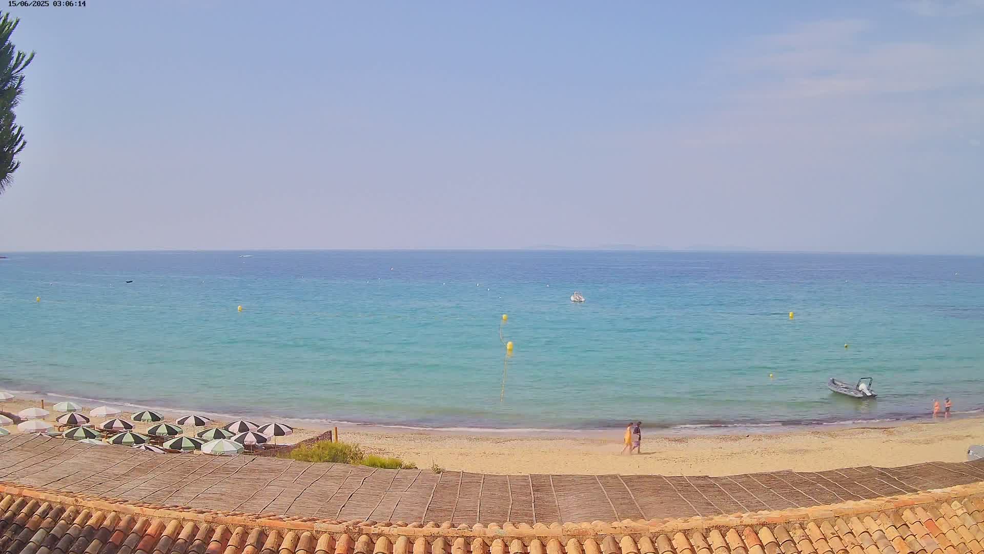A sandy beach with turquoise water, several beach umbrellas, and a few people walking on the sand under a clear, sunny sky.