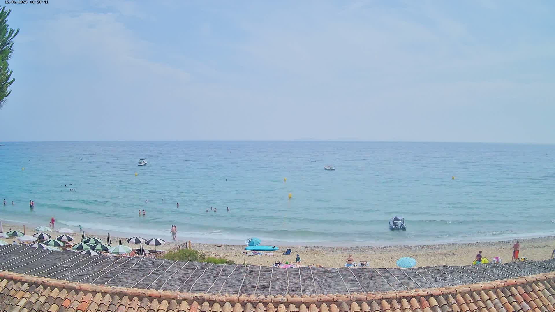 A sandy beach with people swimming and relaxing under umbrellas, overlooks a calm, light blue sea with several small boats on a partly cloudy day.
