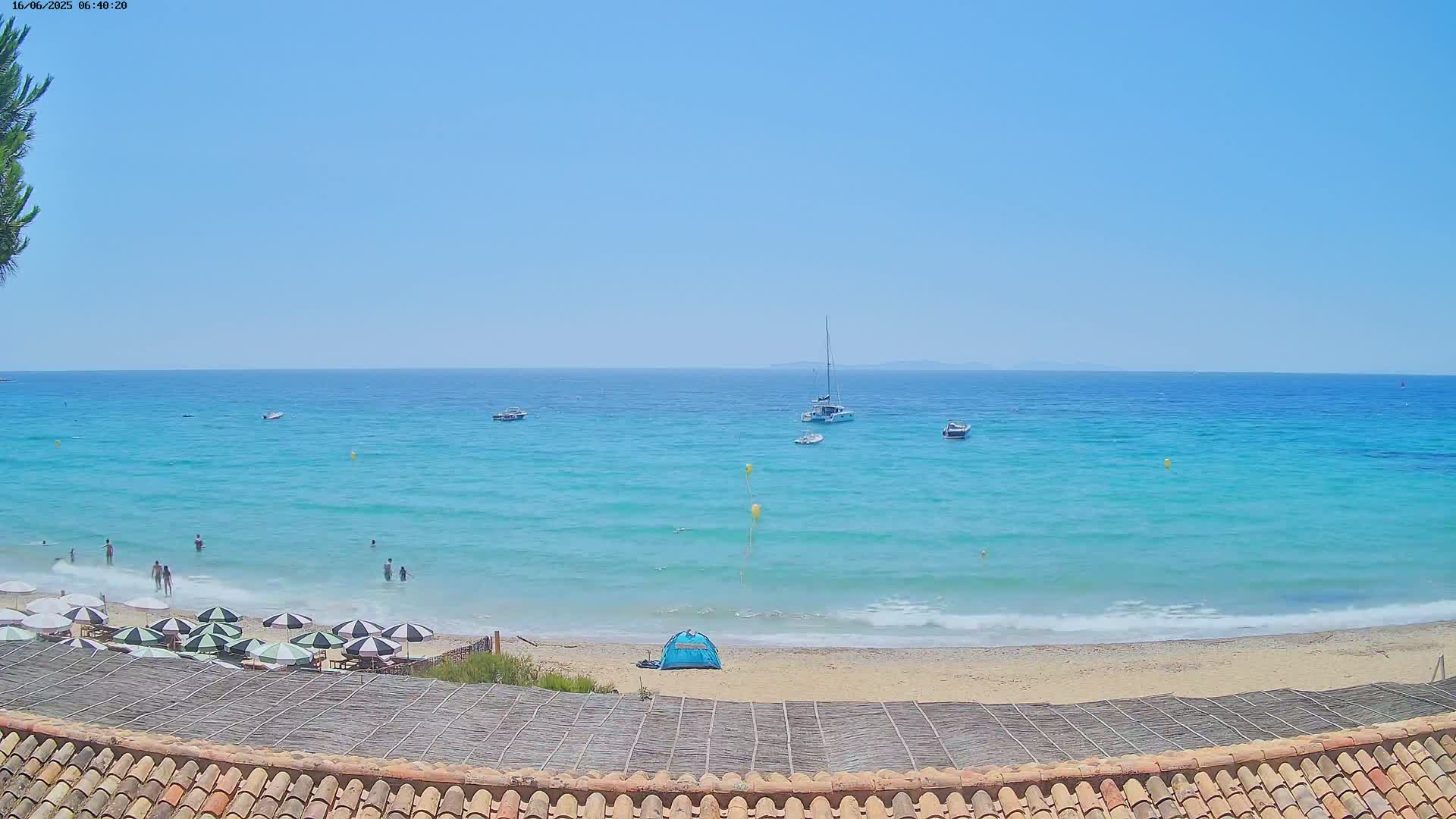 A sandy beach with turquoise water and several boats offshore on a sunny day, with people swimming and sunbathing, and umbrellas set up on the sand.