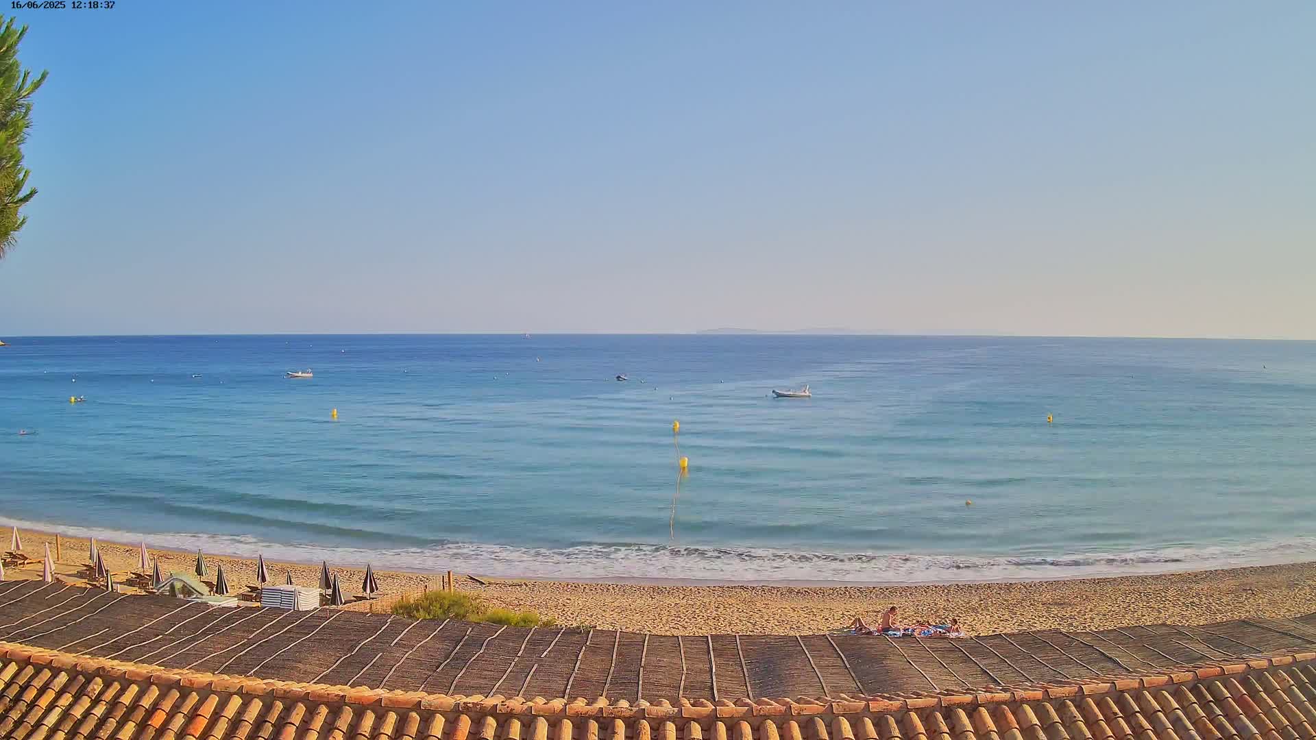 A sandy beach with calm, light-blue ocean, a few boats, and several people relaxing under beach umbrellas on a sunny day.