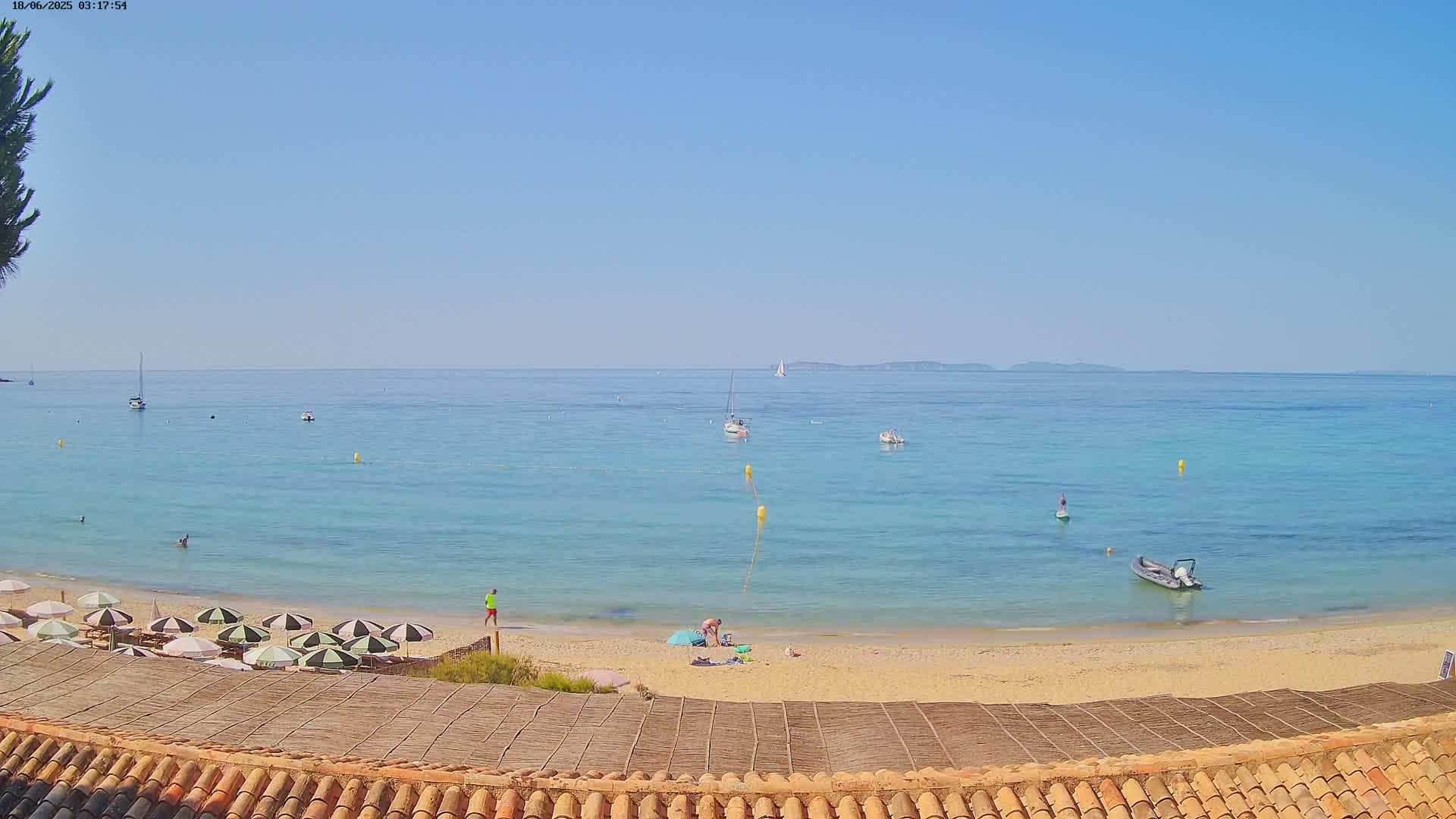 A sandy beach with striped umbrellas and several small boats in a calm, light-blue sea under a clear sky.