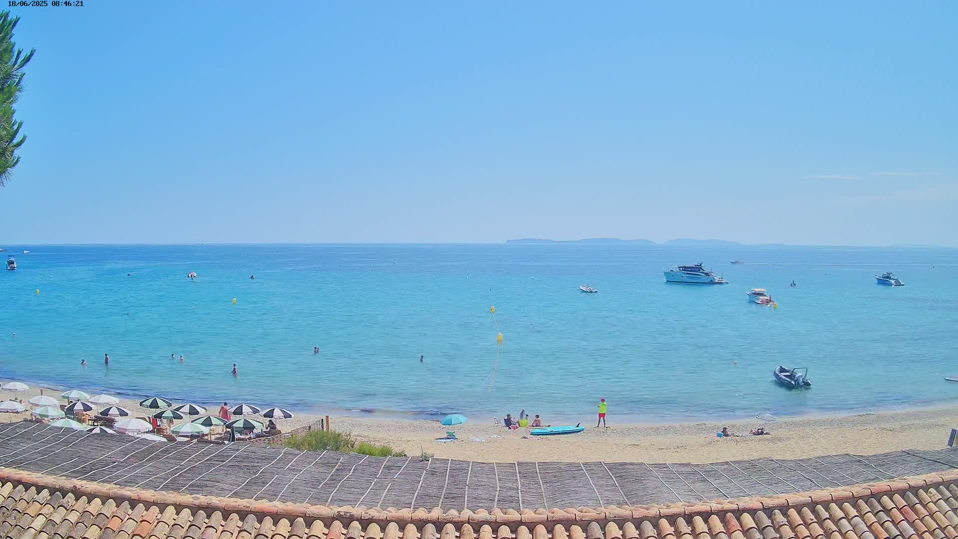 A sandy beach with several people and umbrellas is seen from above, overlooking a calm, turquoise ocean with boats and a clear blue sky.