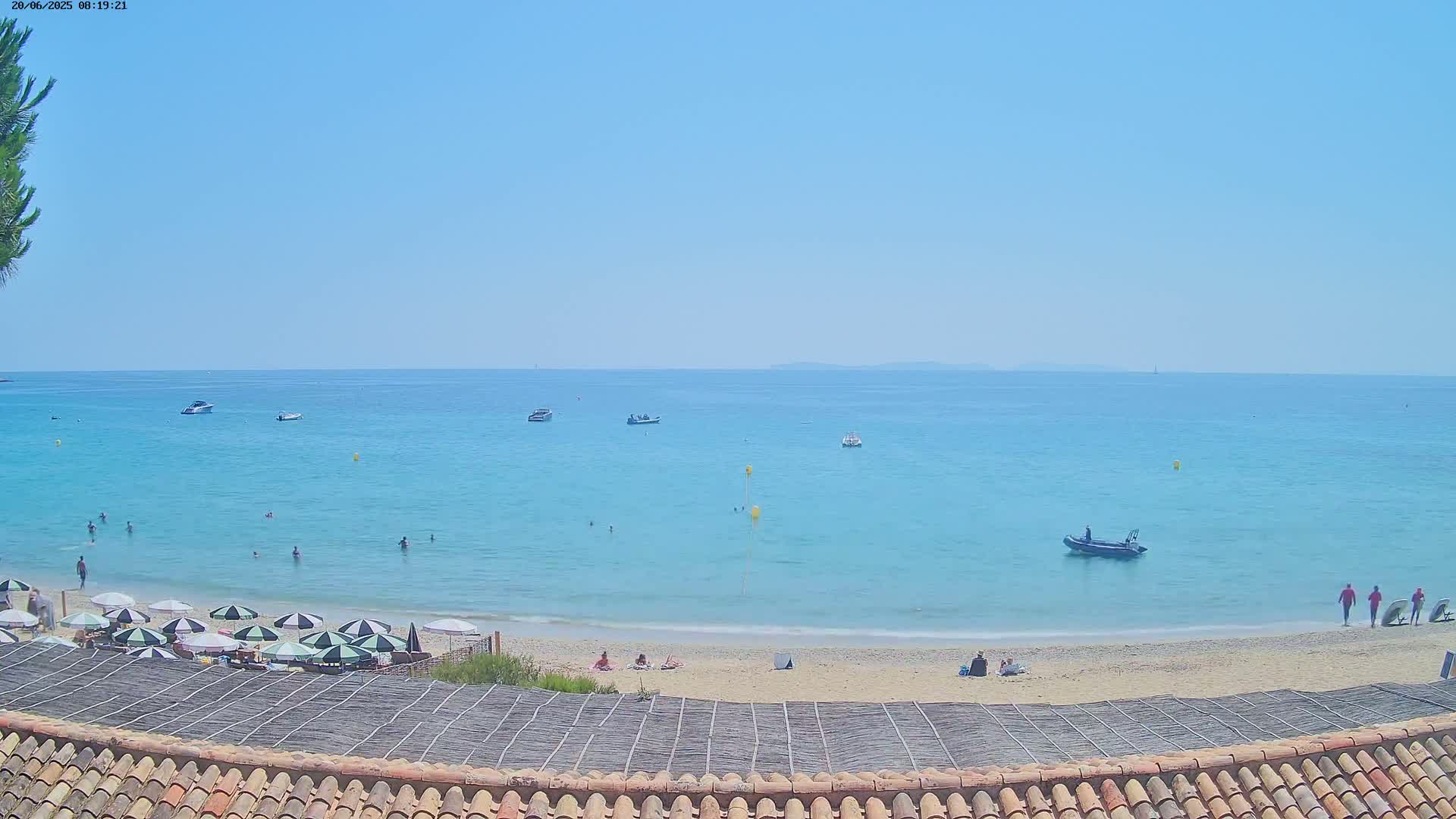 A sandy beach with several people and umbrellas is visible, overlooking a calm, light-blue ocean with a few boats and people swimming under a clear, bright sky.