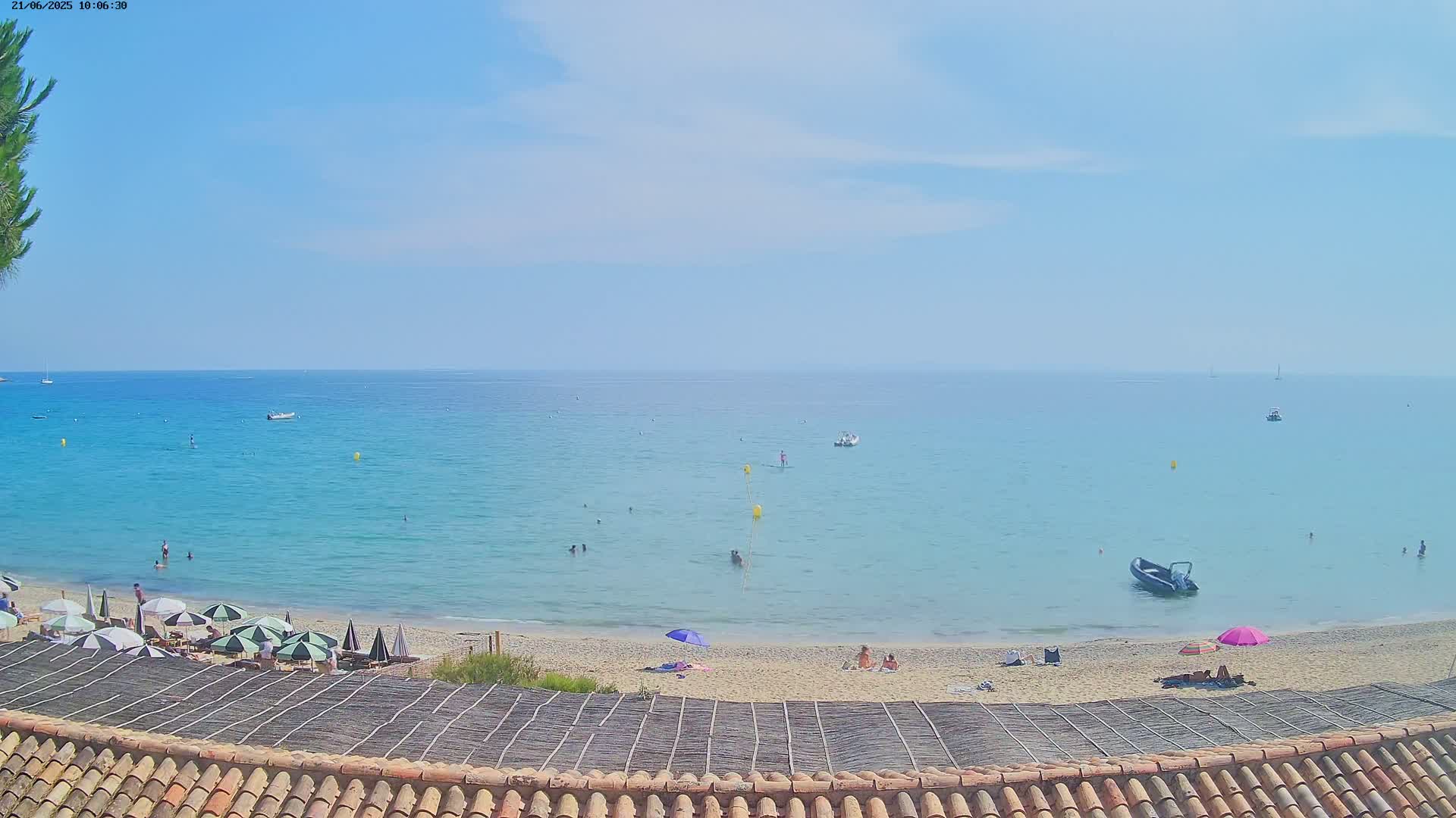 A sandy beach with several people and umbrellas under a mostly clear sky overlooks a calm, light-blue sea with several small boats.