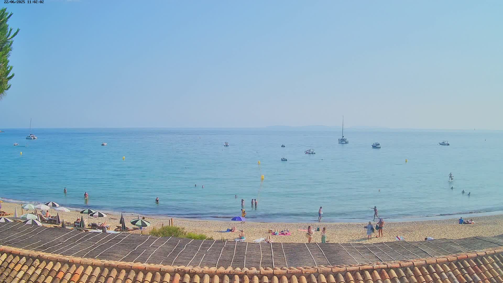 A sandy beach with people sunbathing and swimming in calm, clear blue water, under a bright, sunny sky, with several boats visible in the distance.