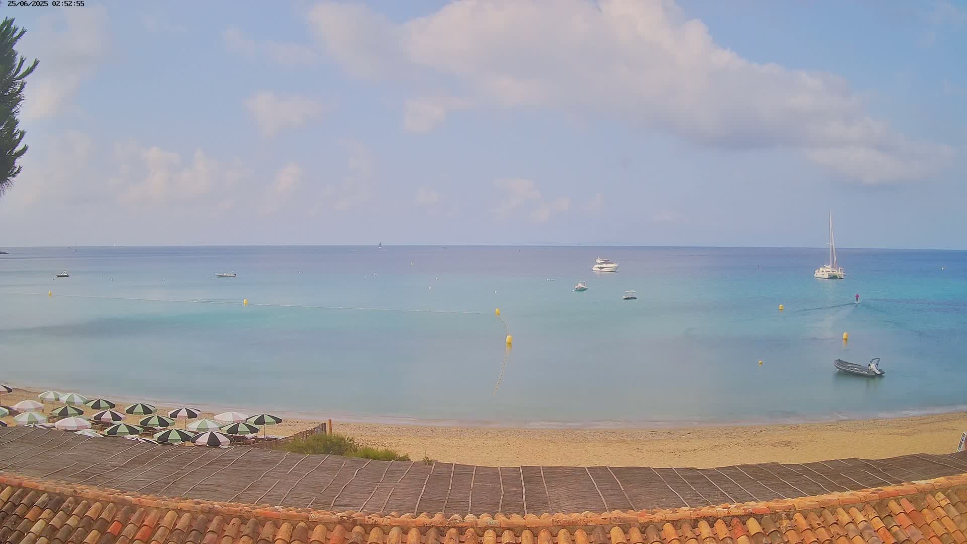 A sandy beach with several closed umbrellas is visible below a calm, light-blue sea with several boats under a partly cloudy sky.