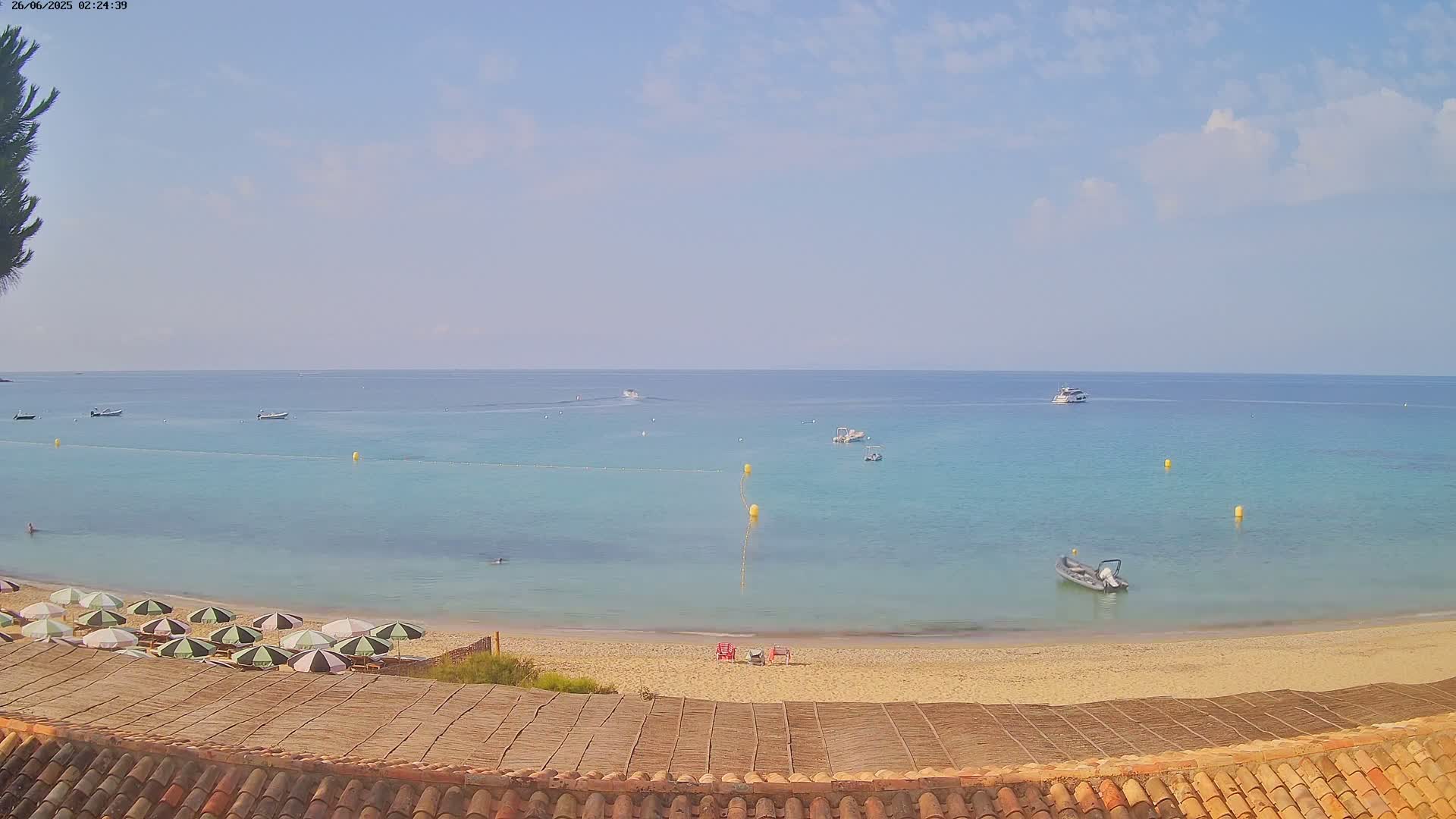 A sandy beach with turquoise water, several small boats, and a row of striped umbrellas under a mostly clear sky.
