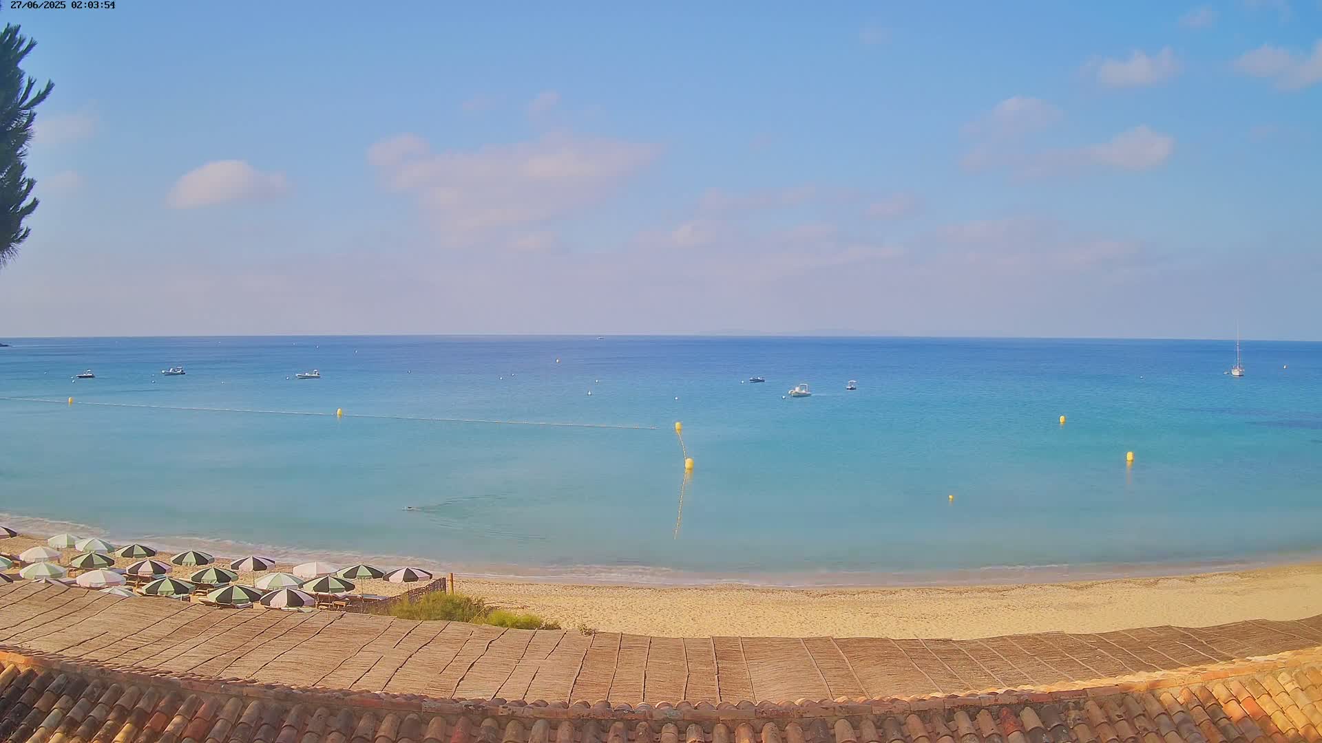 A sandy beach with green and white striped umbrellas is overlooked by a tiled roof under a mostly sunny sky, with calm, turquoise water and several boats visible in the distance.