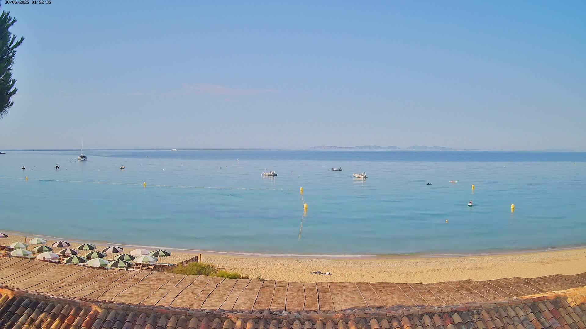 A sandy beach with several closed umbrellas is visible from a tiled roof overlooking calm, light-blue water with boats and buoys, under a clear, sunny sky.