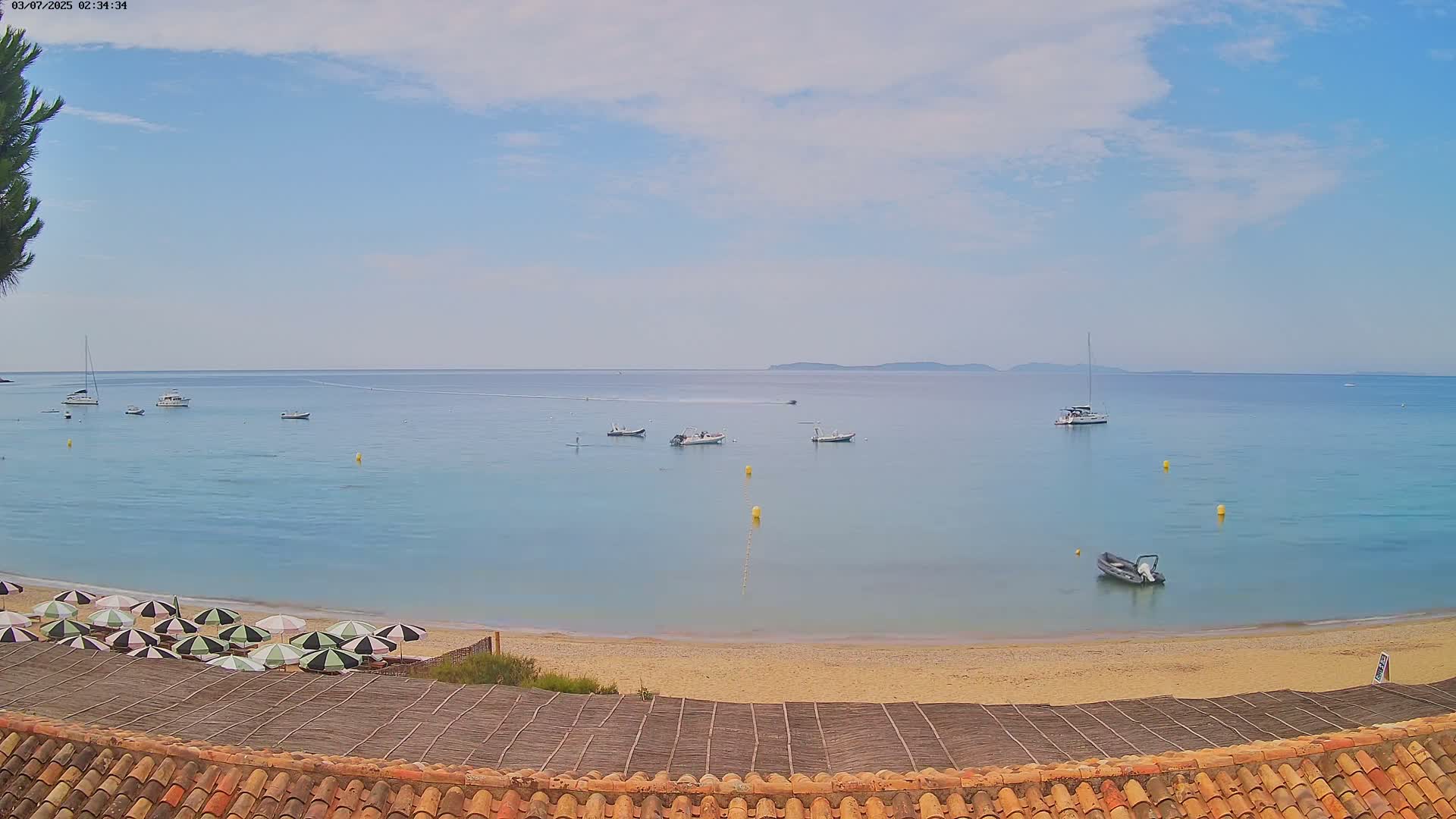 A sandy beach with several boats in the calm, blue water under a partly cloudy sky is seen from above a tiled roof.