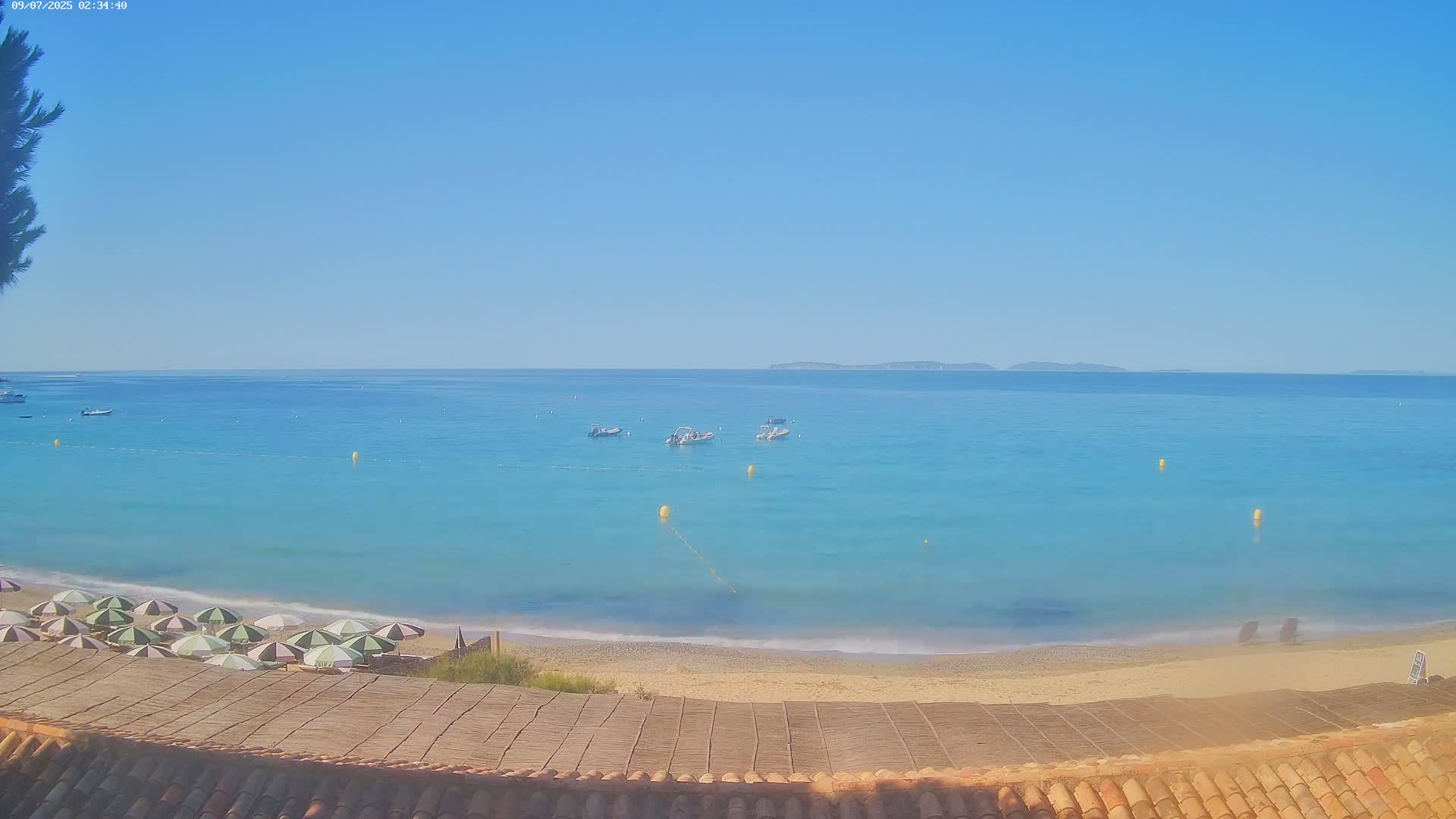A sandy beach with several closed umbrellas overlooks a calm, clear turquoise sea with a few small boats and distant islands under a bright blue sky.
