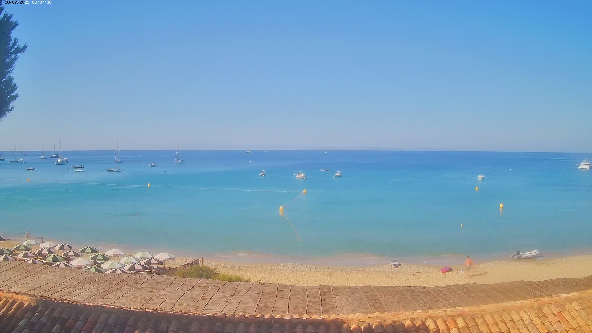 A sandy beach with turquoise water and several boats offshore on a clear, sunny day, viewed from above a thatched roof.