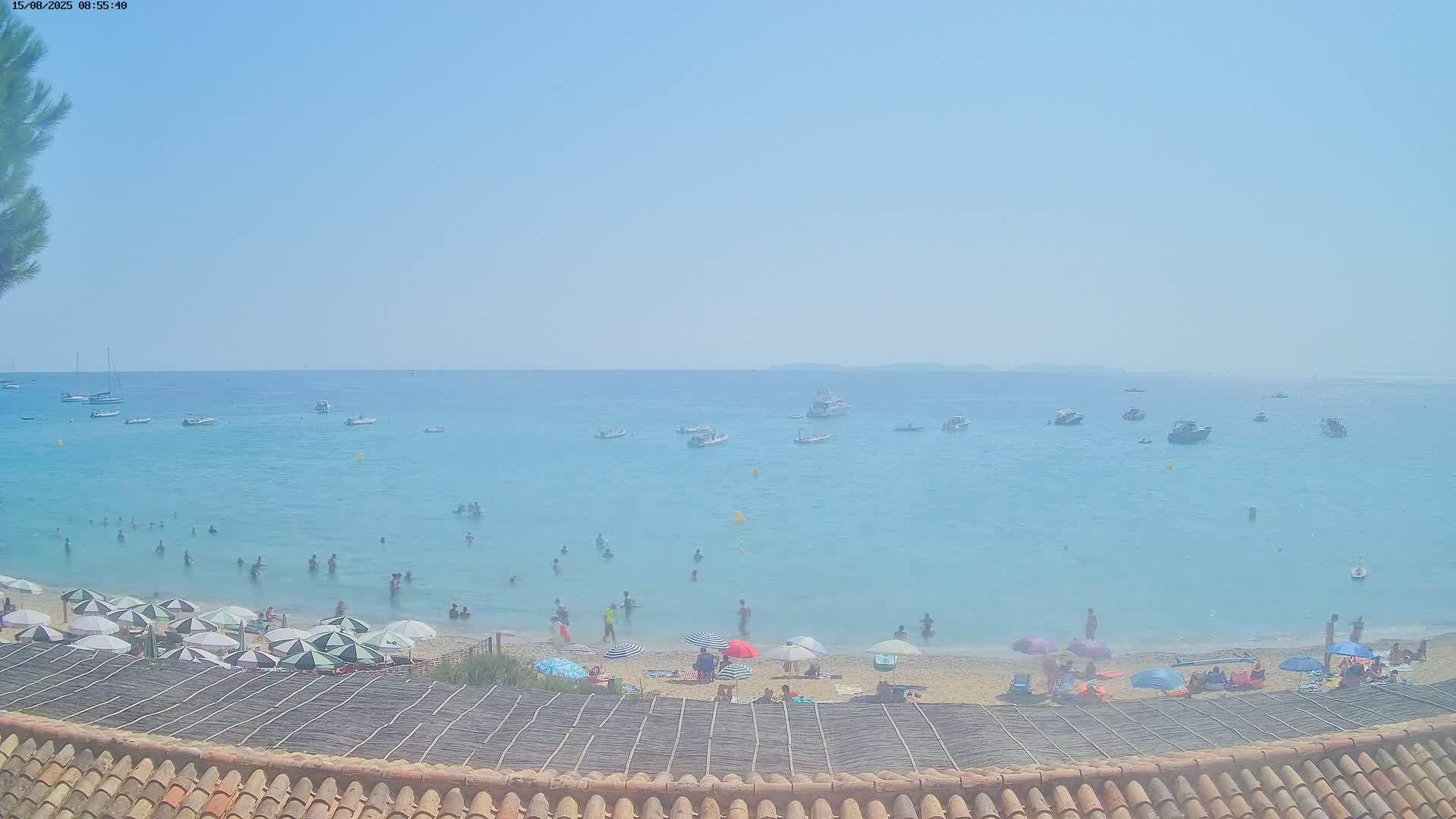 A sandy beach with turquoise water and several boats offshore on a clear, sunny day, viewed from above a thatched roof.