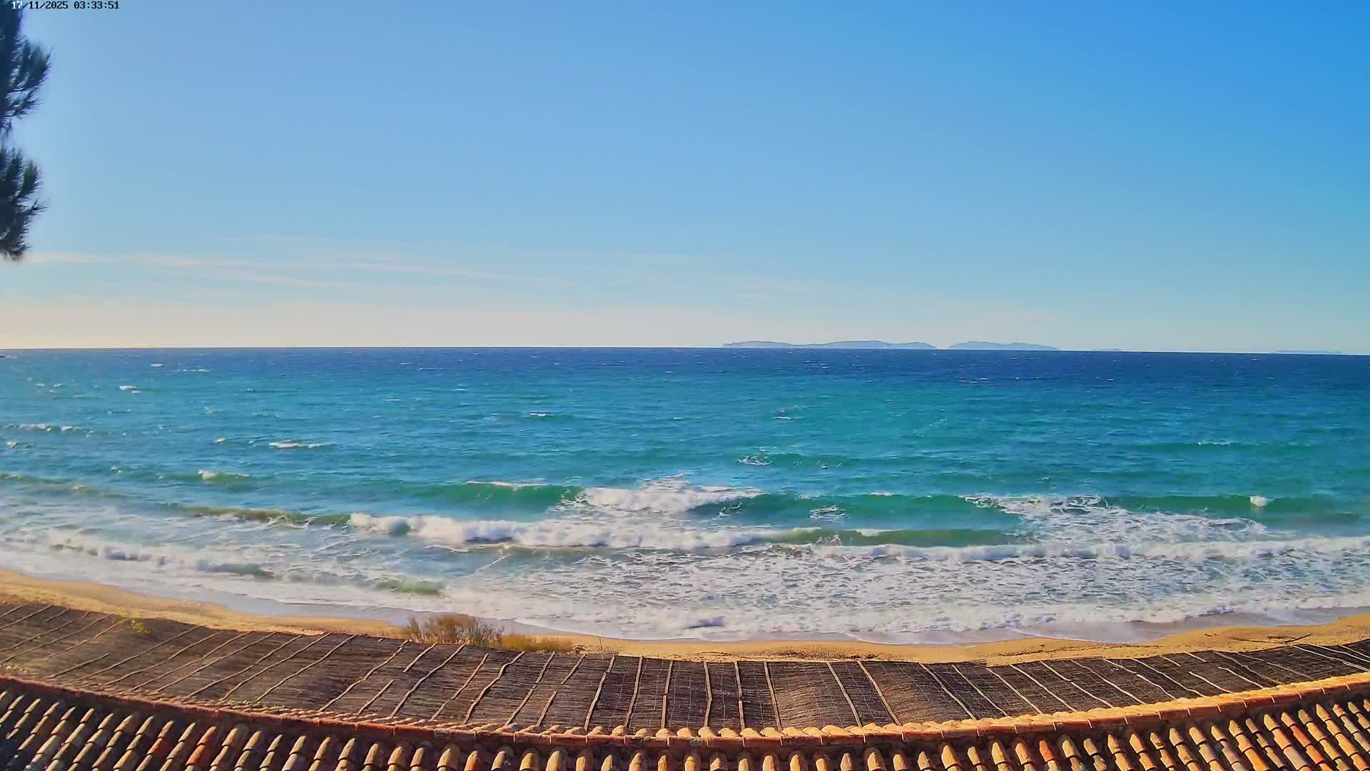 On a sunny day with a clear blue sky, a tiled roof frames a view of a sandy beach where turquoise ocean waves break, with distant islands visible on the horizon.