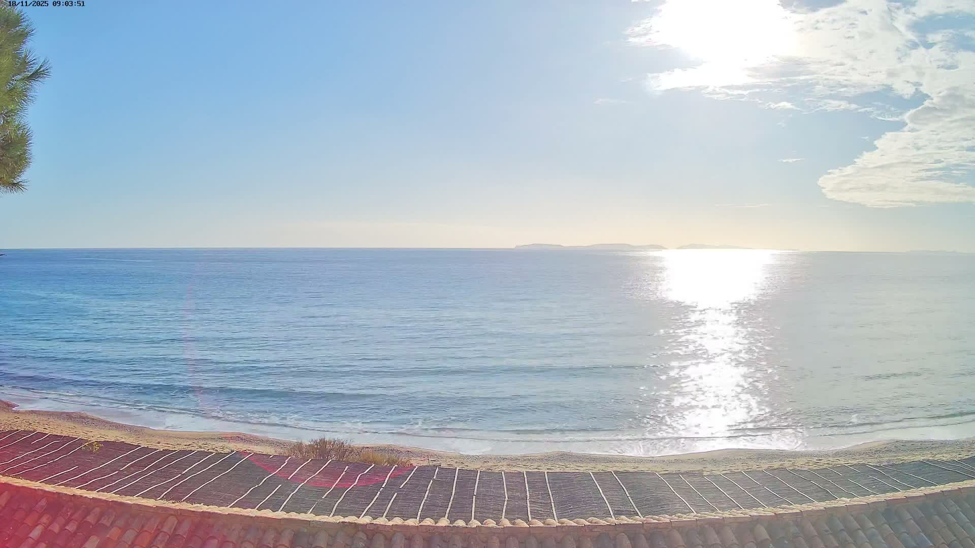 On a sunny day with a clear blue sky, a tiled roof frames a view of a sandy beach where turquoise ocean waves break, with distant islands visible on the horizon.
