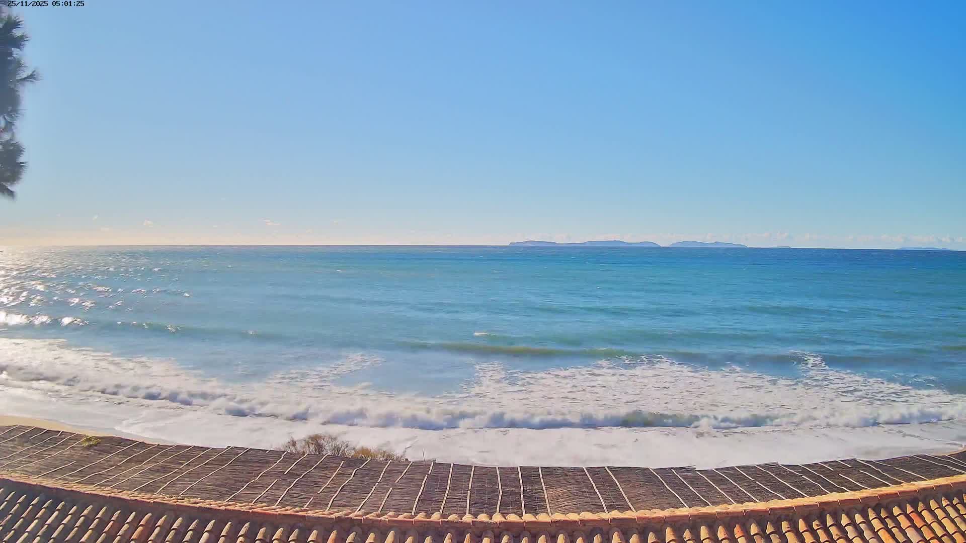 The scene captures a clear, sunny day over a vast blue ocean with white-capped waves breaking on a sandy beach, viewed over a tiled rooftop, with distant land visible on the horizon.