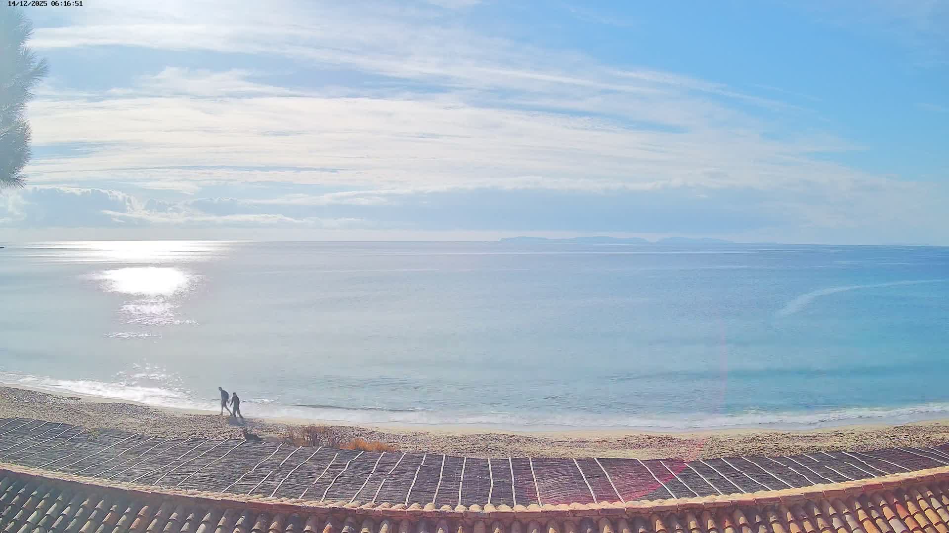 The scene captures a clear, sunny day over a vast blue ocean with white-capped waves breaking on a sandy beach, viewed over a tiled rooftop, with distant land visible on the horizon.
