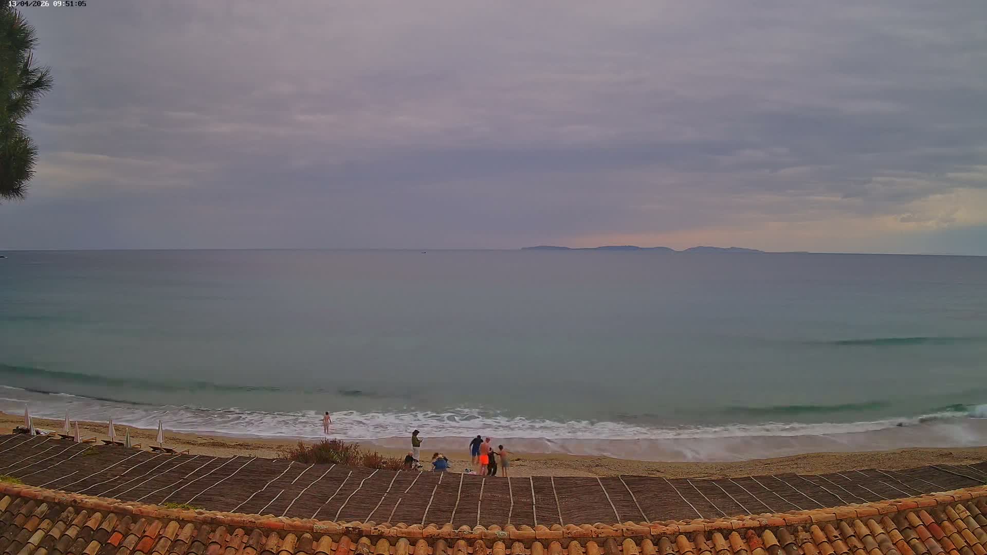 The scene captures a clear, sunny day over a vast blue ocean with white-capped waves breaking on a sandy beach, viewed over a tiled rooftop, with distant land visible on the horizon.