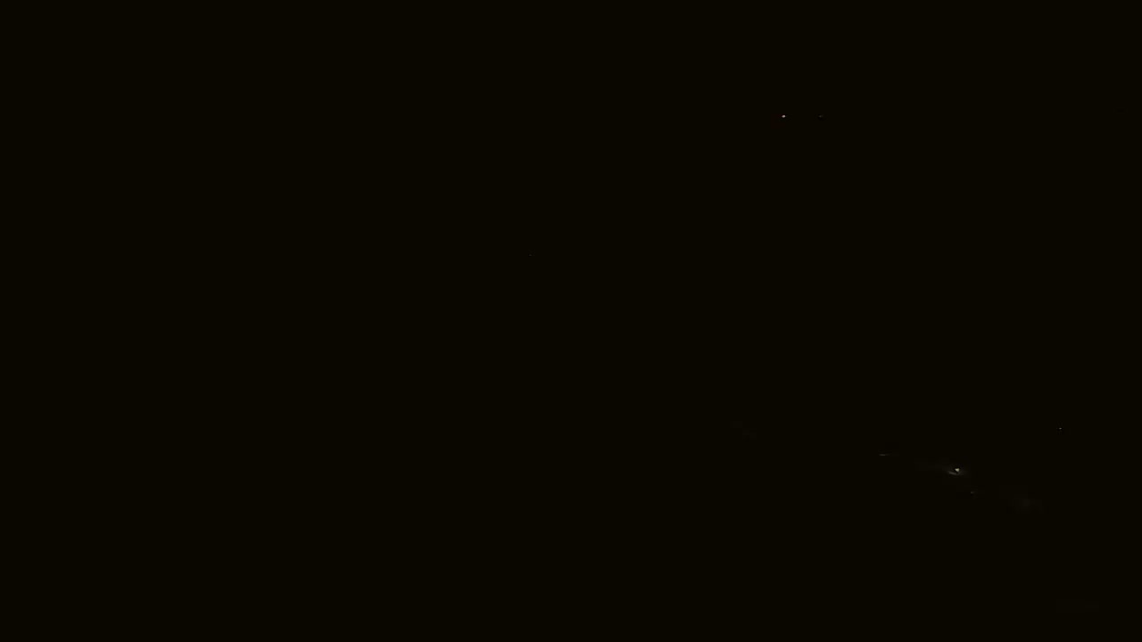 A nighttime long shot shows a few faint lights on the dark ground, and a couple of brighter lights in the dark sky.