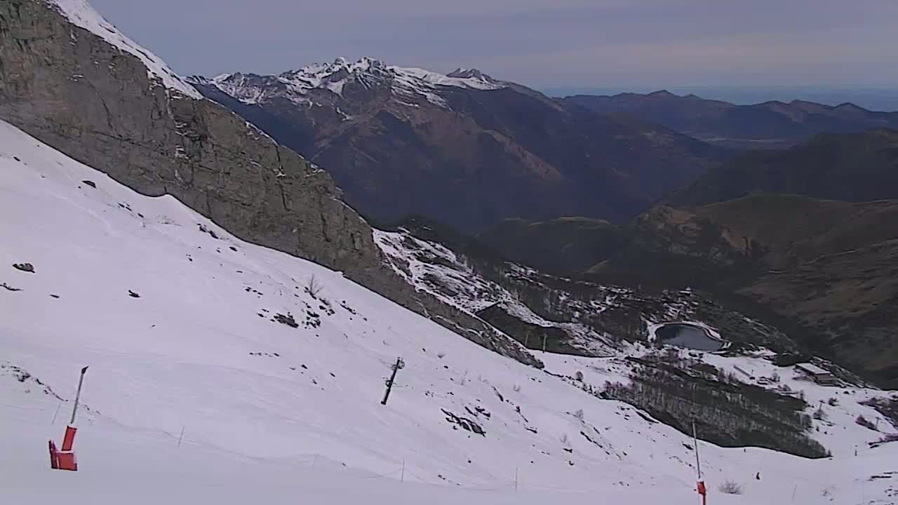 A snow-covered mountain slope descends towards a valley with a small lake, under a mostly cloudy sky.