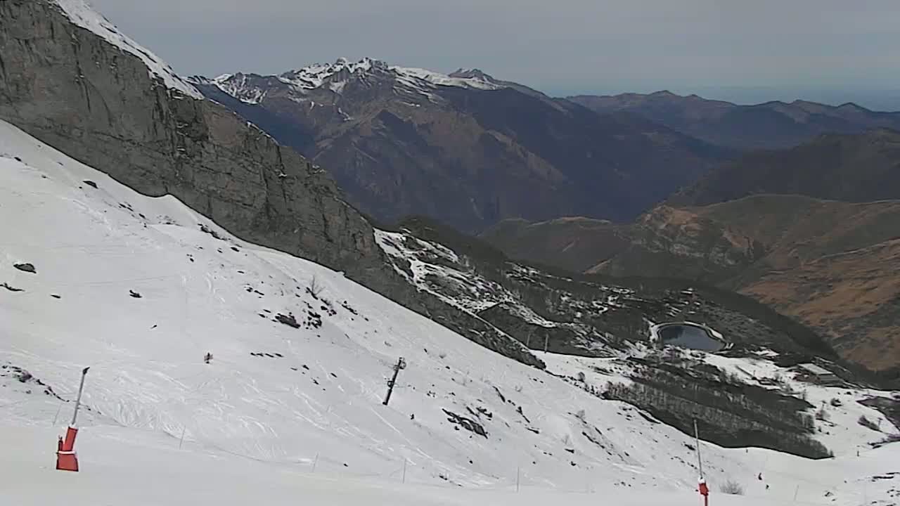 A snow-covered mountain slope with ski runs and lift equipment overlooks a valley of brown and green mountains under a cloudy sky.