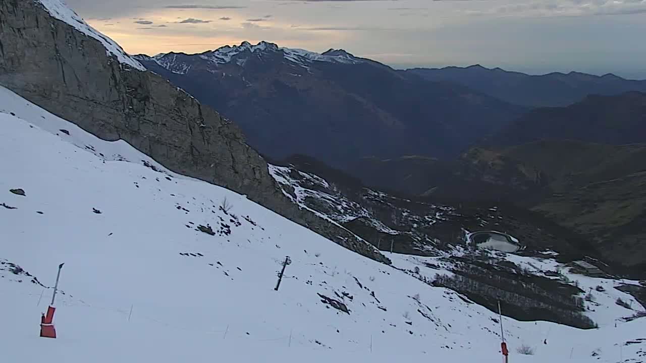 A snow-covered mountain slope with ski markers descends towards a valley, under a partly cloudy sky at dawn, overlooking distant mountain ranges.