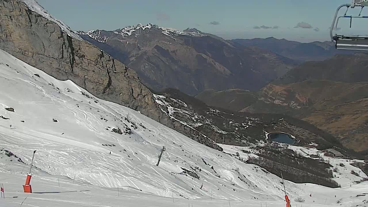 A snow-covered mountain slope with ski tracks, overlooking a valley and distant mountains under a clear sky.