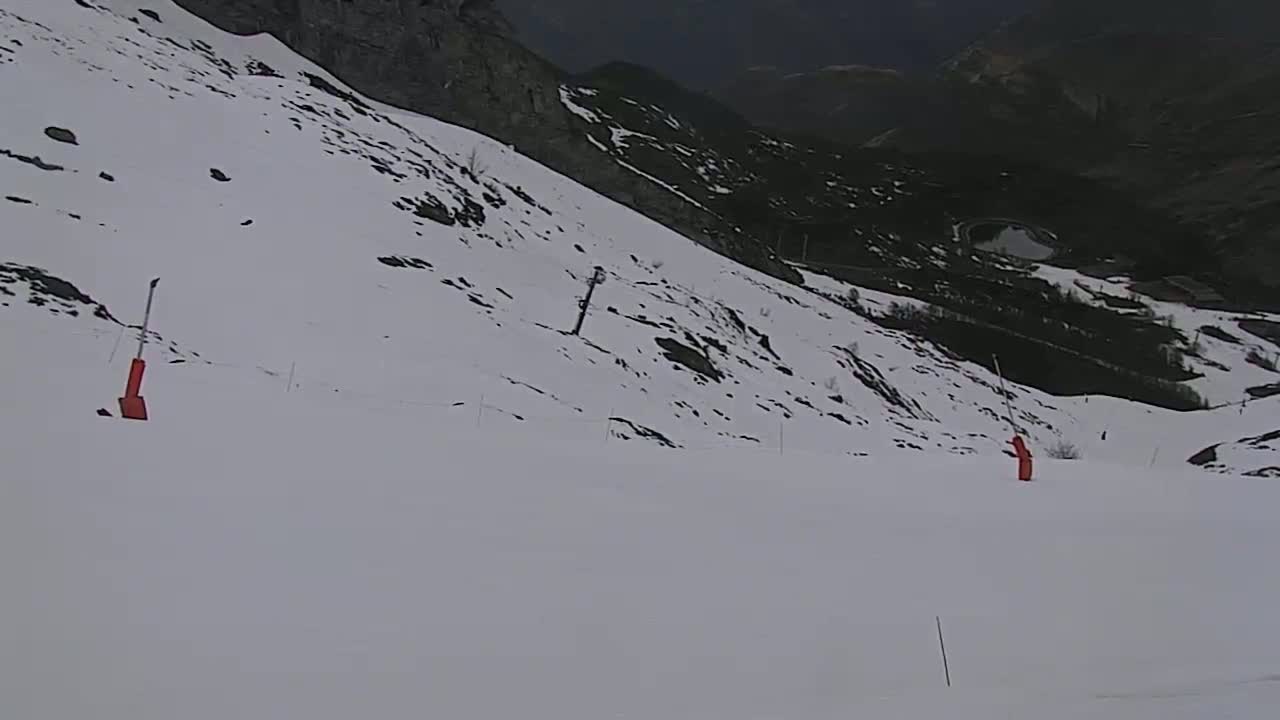 A snow-covered mountain slope with sparse vegetation and red markers, overlooking a valley below on an overcast day.