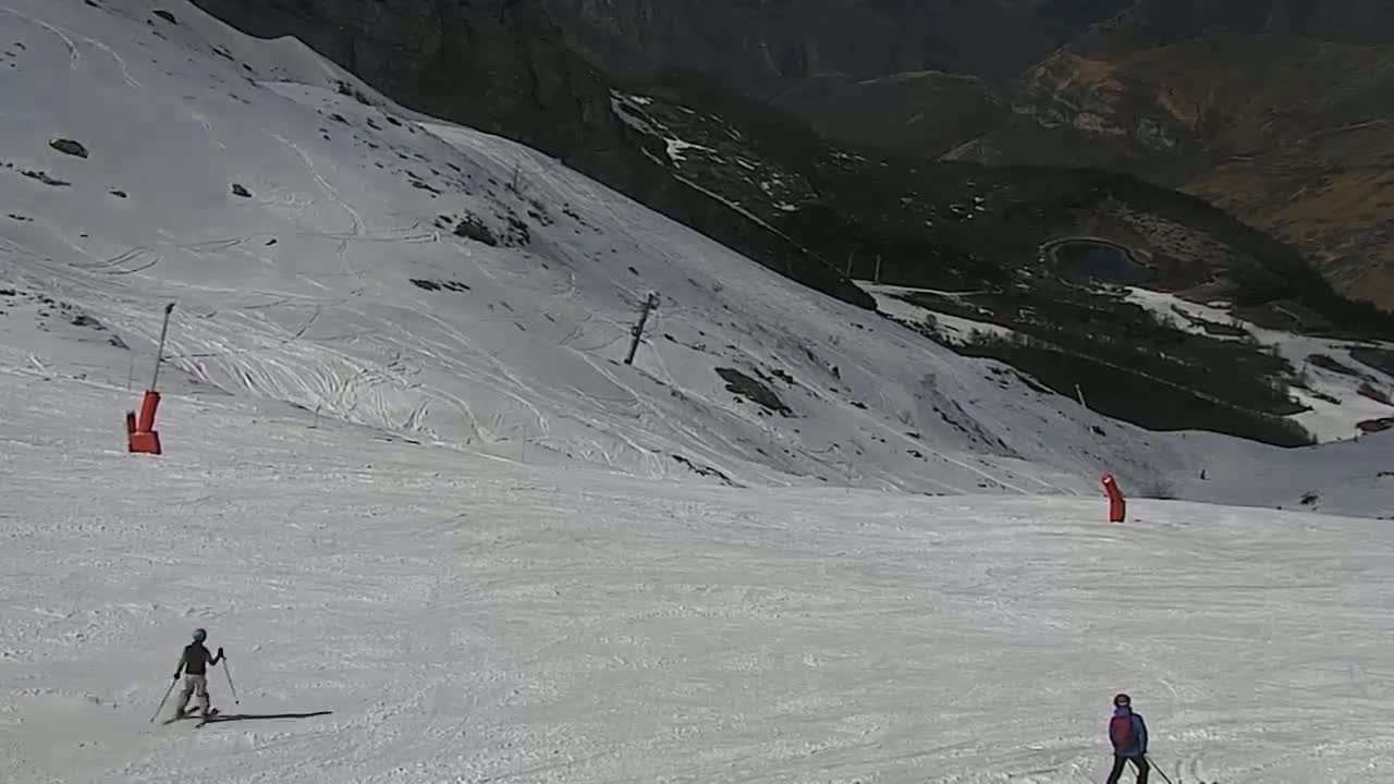 A snowy ski slope with two skiers and two orange markers is visible, with a mountainous landscape in the background under sunny conditions.