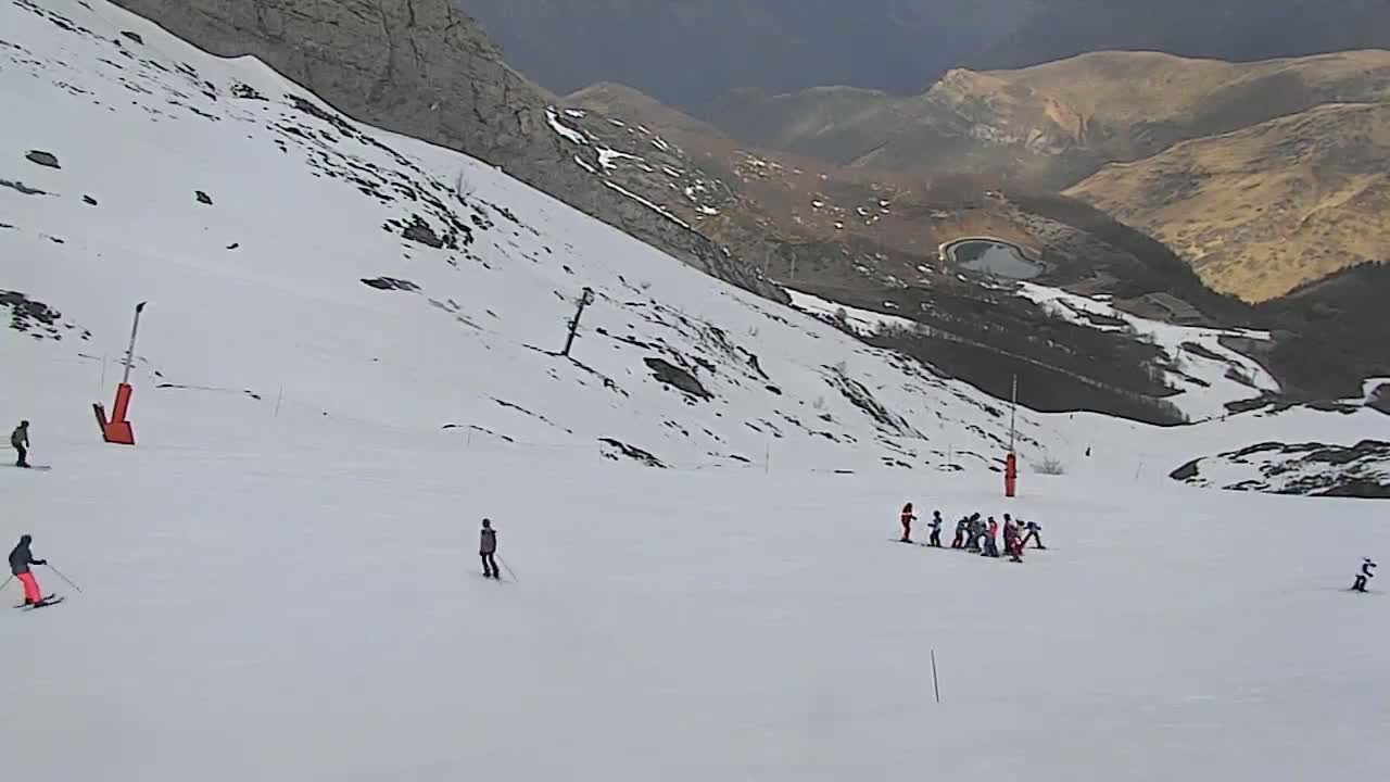 A snowy mountain slope with several skiers and snowboarders on a mostly clear day, with a mountain range visible in the background.