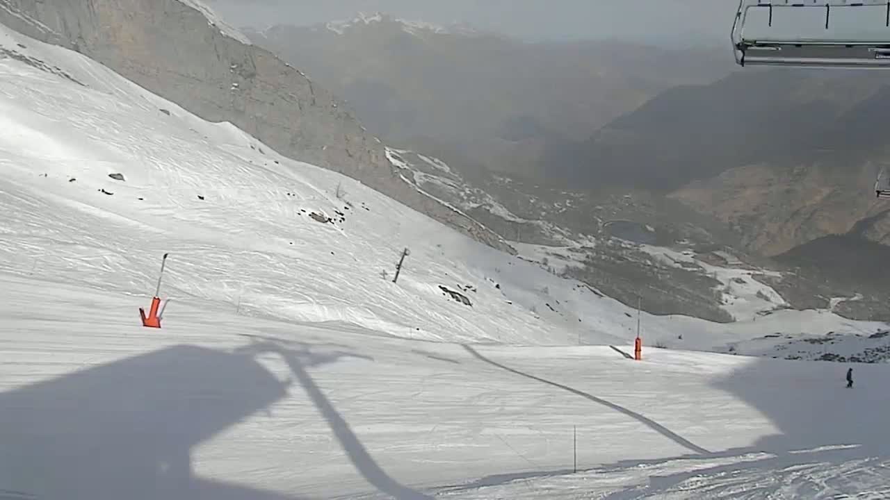 A snowy mountain slope with a hazy view of distant mountains, showing a ski run with two orange markers and the shadows of a ski lift.