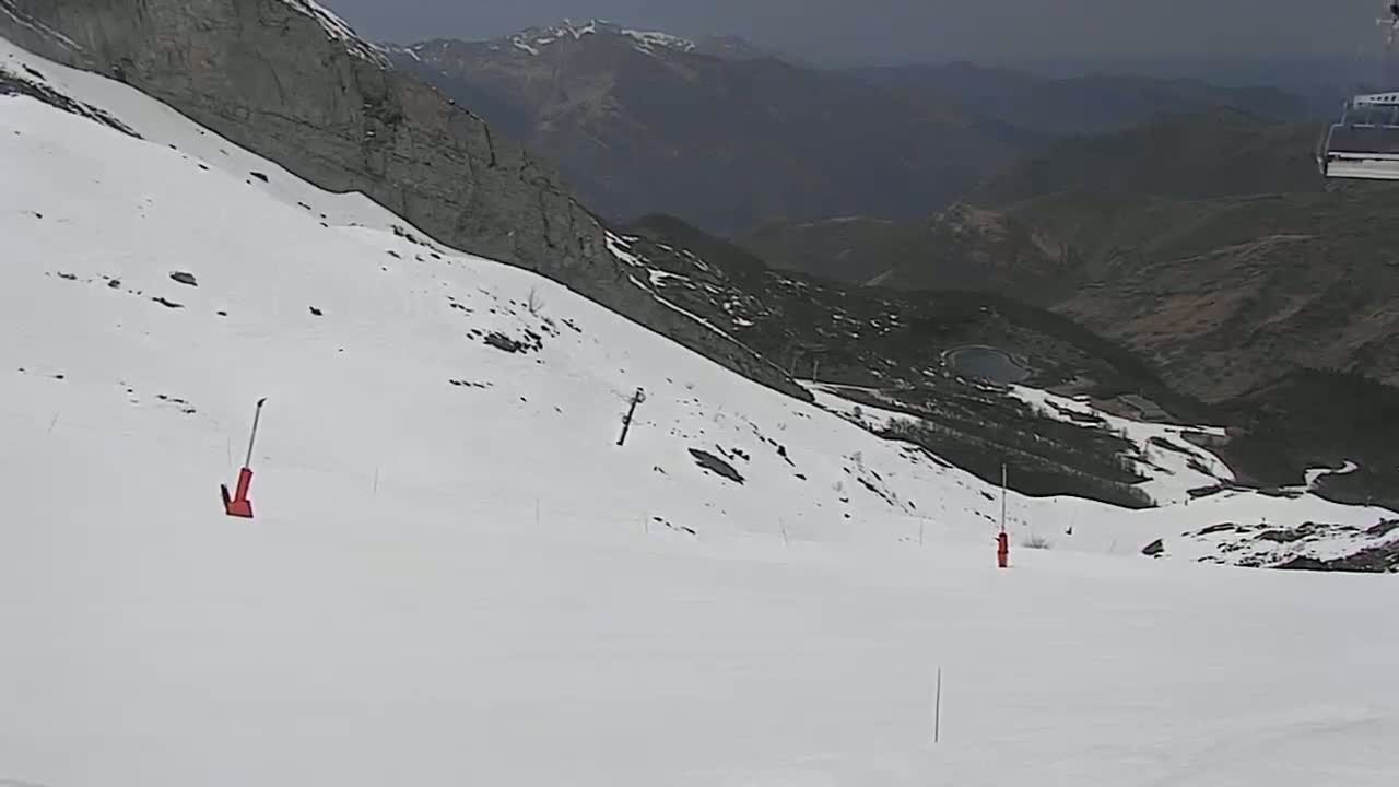 A snow-covered ski slope descends towards a mountain valley under an overcast sky.