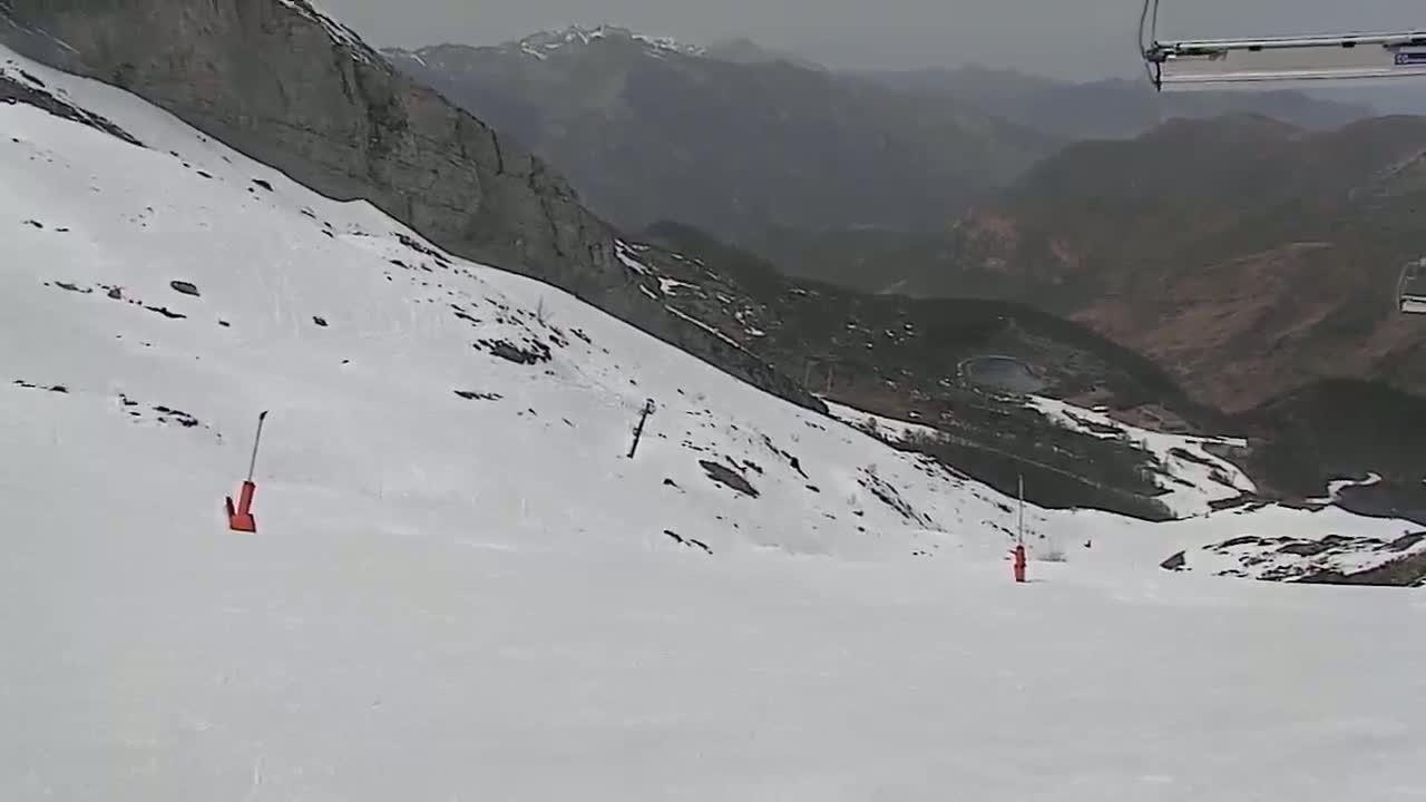 A snowy mountain slope with two orange markers is visible, overlooking a valley under an overcast sky.